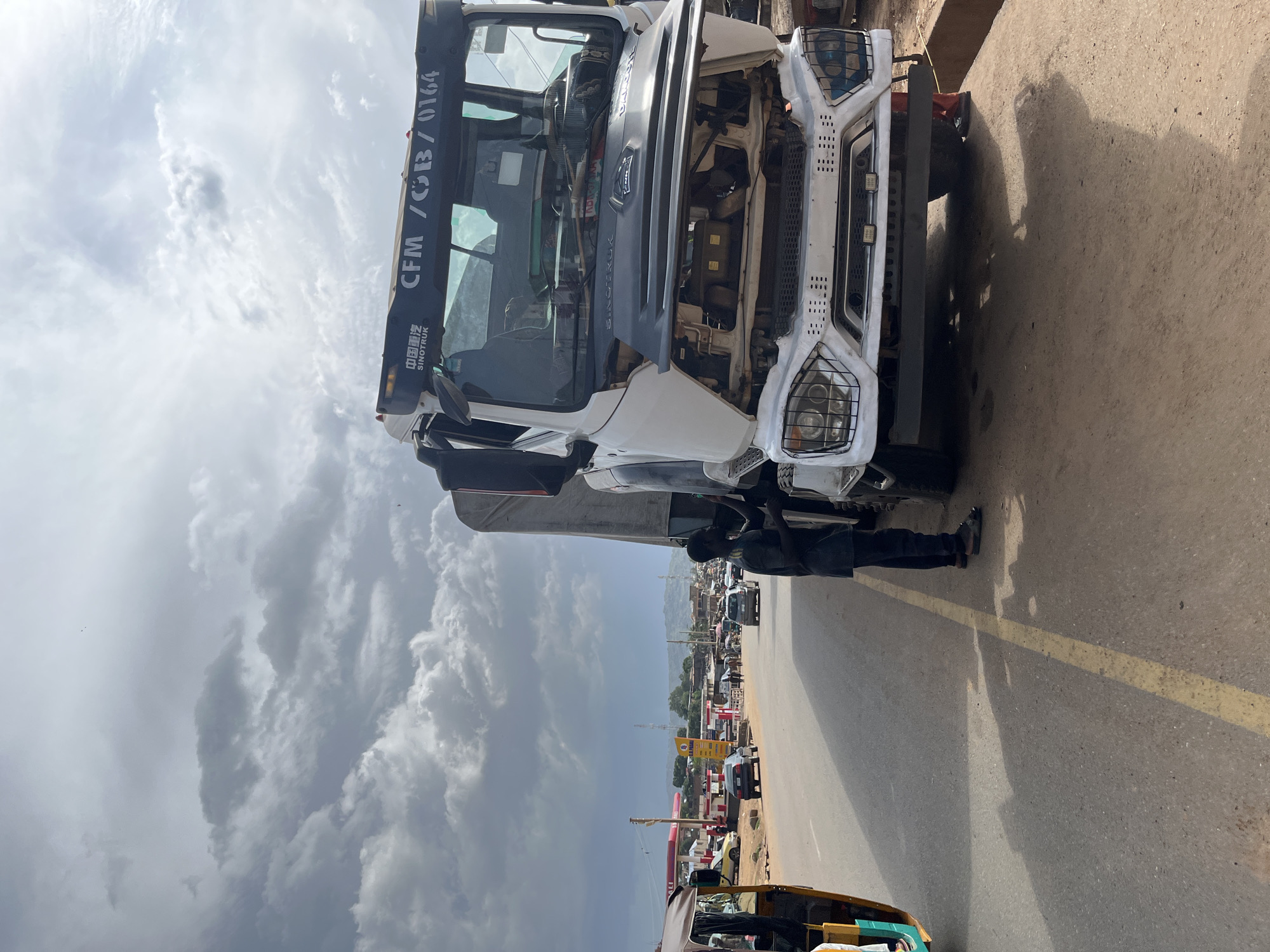 The image captures a roadside scene in Jos, Nigeria. A large white truck, possibly a Sinotruk model indicated by the lettering "SINOTRUK" and "CFM / GB / 0164" on its front, has its hood open, suggesting a mechanical issue. A Black man, dressed in a dark shirt and pants, is standing next to the truck, appearing to be working on it. The truck is positioned on the right side of a paved road, with its front tires visible.
In the foreground on the left, a three-wheeled auto-rickshaw, commonly known as a "Keke Napep" in Nigeria, is partially visible. The rickshaw has a yellow canopy and a person is seated inside. The road is a two-lane asphalt surface with a visible yellow lane marker.
The background reveals a typical urban environment in Jos. There are buildings, including what appears to be a petrol station with red and white pumps, and several shops. A few other vehicles, including cars and a motorcycle, are present on the road, indicating traffic. The sky is partly cloudy with dramatic, dark clouds suggesting an approaching storm or the aftermath of one. The overall atmosphere suggests a functional, albeit potentially interrupted, day on a busy road in Jos.