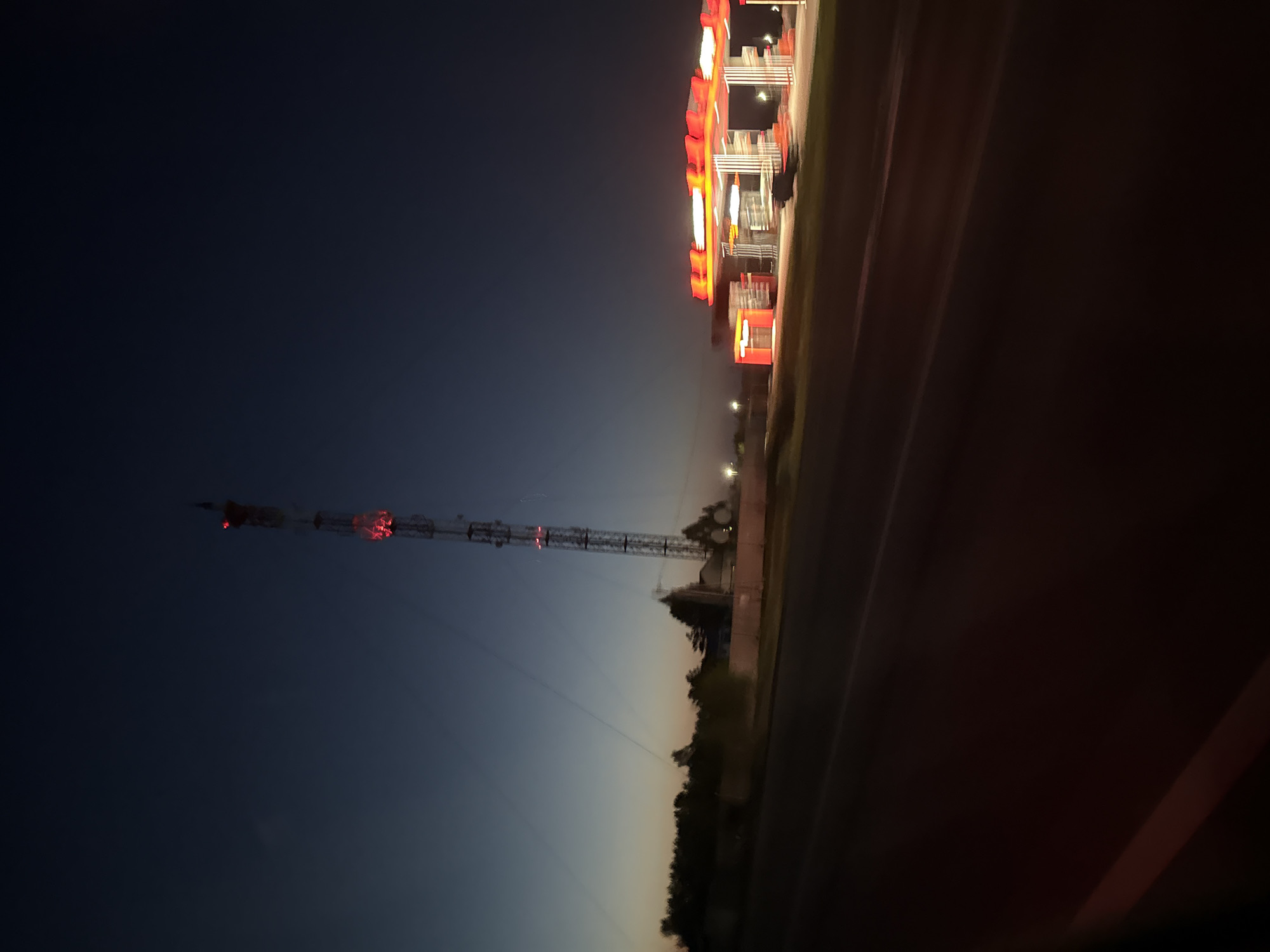 At night, a tall communication tower with red lights stands in an open space adjacent to a line of trees. To the right, a brightly lit building with vertical rows of red and white lights is partially visible. The location appears to be near a road.