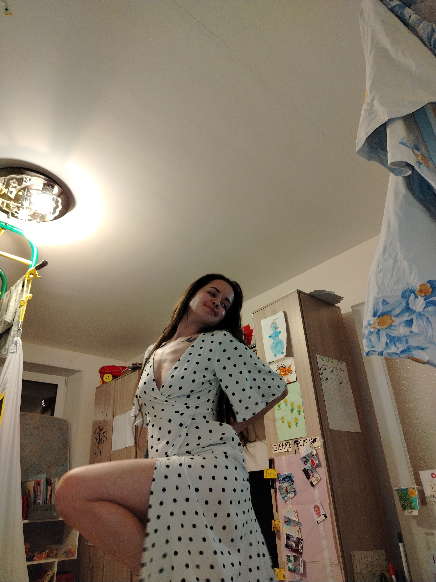 A woman stands in a room with a low-angle perspective. She is fair-skinned and wears a white, knee-length dress with black polka dots, featuring a V-neck. Her long, dark hair is loose.
The room includes a ceiling light with decorative metalwork. Adjacent to the woman on the left is a child's indoor swing or play area with colorful framing and white fabric. A small bookshelf sits below.
To her right, a tall cabinet is decorated with children's drawings and charts attached to it. A hanging white cloth with blue floral designs is positioned in the top right corner of the frame.
The environment appears domestic and somewhat cluttered, with a mix of personal items and children's belongings scattered around the room. There is no evidence to identify the exact location in the city of Noviy, Russia.