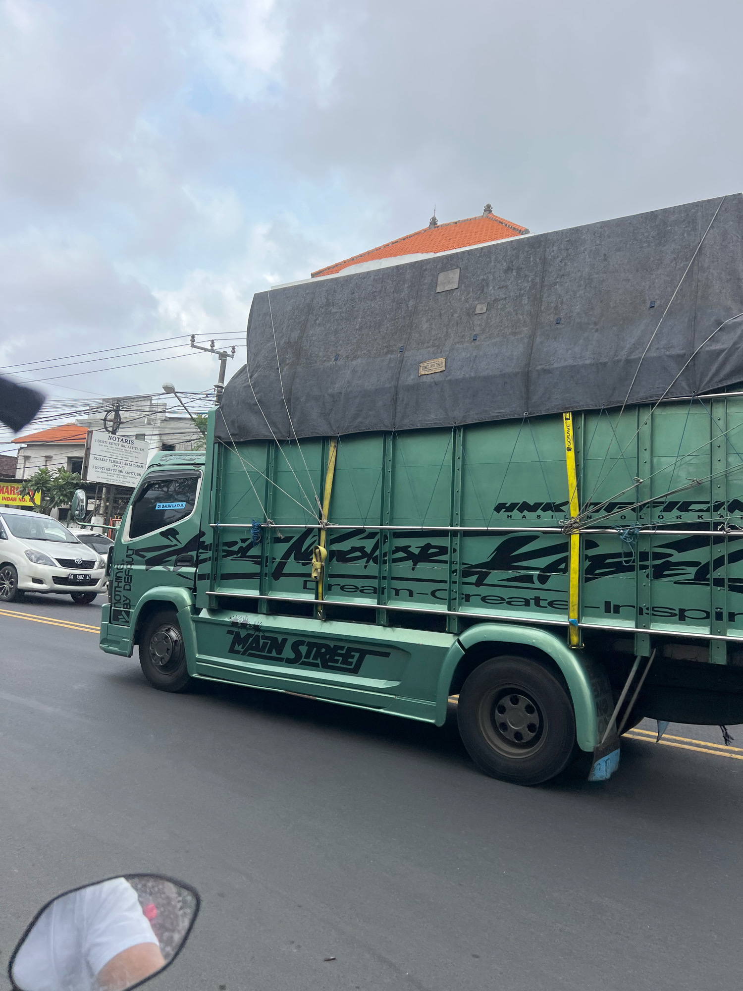 This is a medium shot, eye-level, outdoor, natural light, vertical photograph taken from a vehicle, likely a motorcycle based on the rearview mirror in the foreground. The image captures a light green colored truck with a dark gray tarp covering its cargo area, driving on a paved road in Denpasar, Indonesia. The truck has black decals on its side, including the words "MAIN STREET" and "DREAM-CREATE-INSPIRATION". A white car is visible in the left lane of traffic, and a building with an orange tiled roof and a sign with Indonesian text is in the background. The sky is overcast and the road appears to be wet, suggesting it may have recently rained. Electrical wires and utility poles are also visible along the roadside.