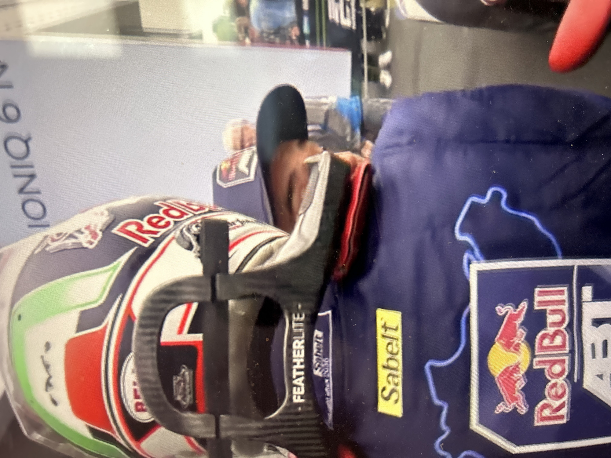 A person is reclining, wearing a blue racing suit with a Red Bull logo, a Sabelt patch, and a Featherlite logo. They are also wearing a black cap. A helmet with a Red Bull logo, green, white, and red stripes is beside them. The background appears to be a bright indoor space. "IONIQ 6" is visible. There is no concrete information in the image to determine the location is in Sofia, Bulgaria.
