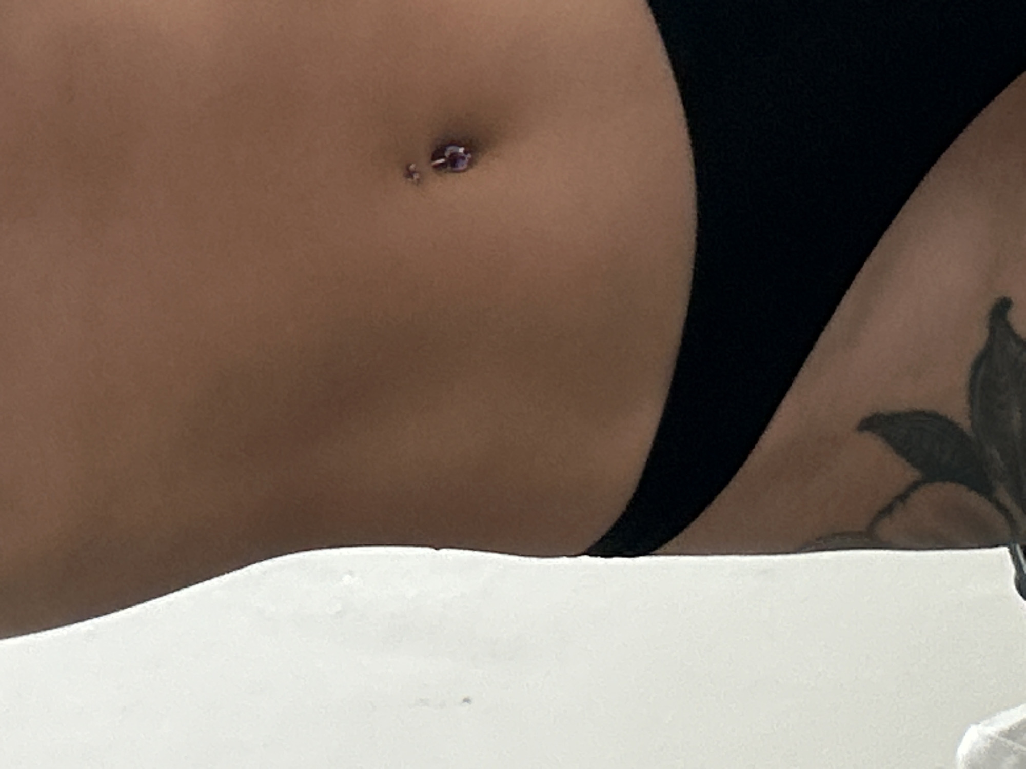 The image displays a close-up, side profile of a person's torso, focusing on the midsection. The skin tone is medium to dark brown. A navel piercing is visible, adorned with a small, purple, jewel-like accessory. The lower part of the image shows the top edge of black underwear. On the left side, a portion of a tattoo, featuring dark, leafy or floral elements, is visible on the hip area. The background is a plain, off-white wall, with a slight shadow cast on it to the left of the subject. The lighting is soft and even.