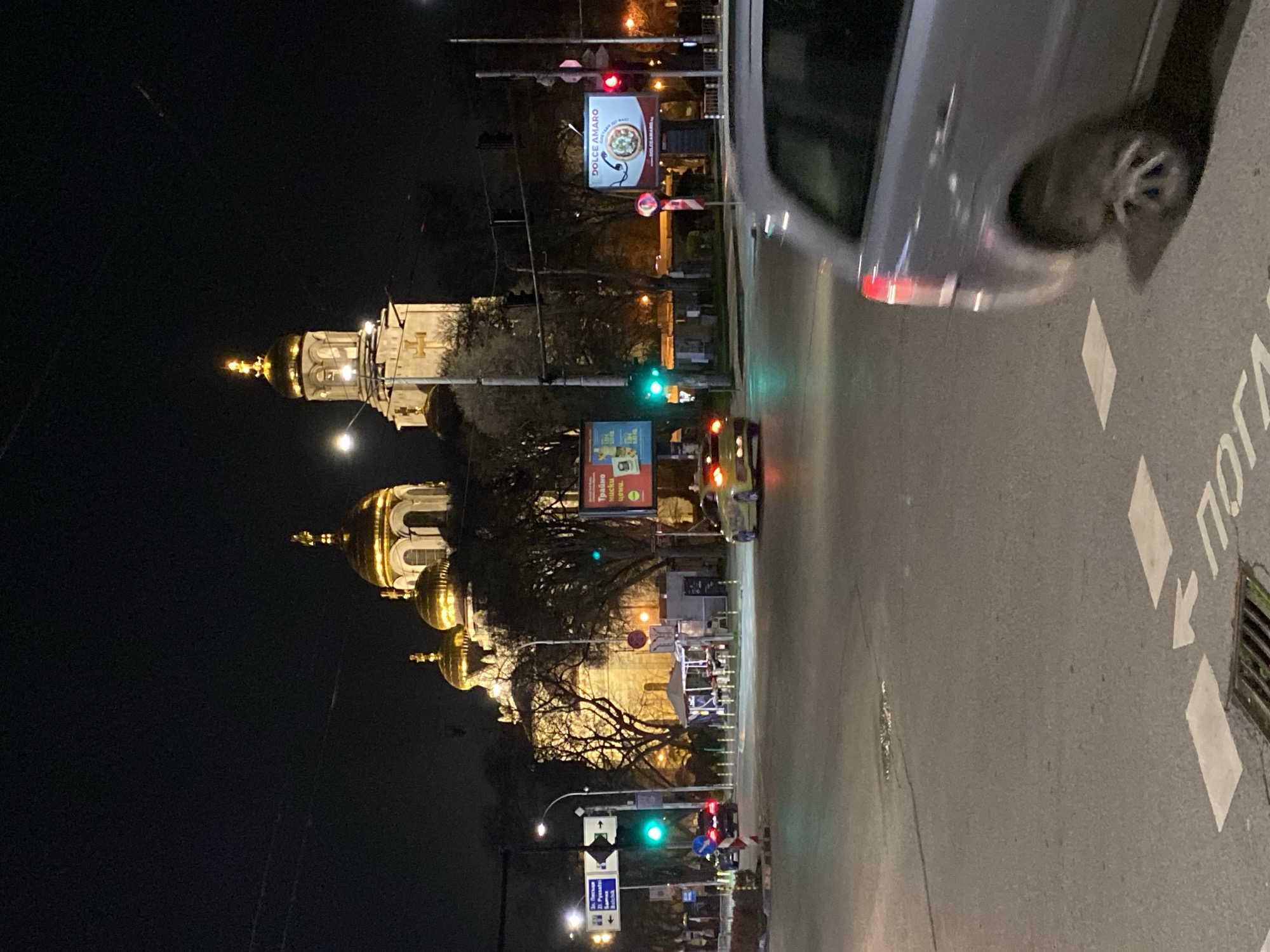 At night, the image shows the Dormition of the Mother of God Cathedral in Varna, Bulgaria, from a street-level perspective. The cathedral, with its illuminated golden domes, stands against the dark sky. Streetlights and traffic signals are visible, with some showing green. A car partially obstructs the upper right side of the frame. The street displays road markings and a drain. Advertising billboards are positioned along the street. Pedestrians can be seen near the traffic lights.
