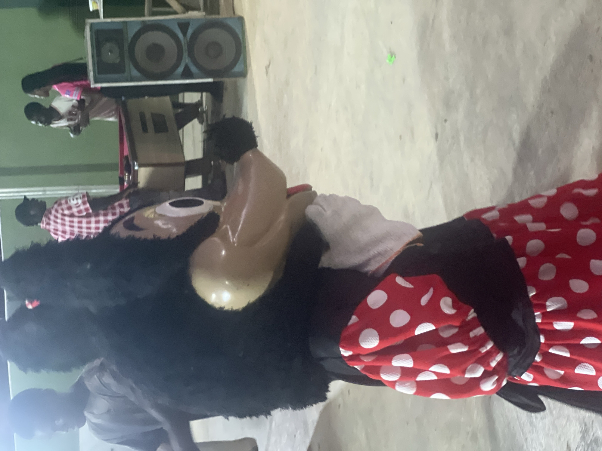 The image shows a Minnie Mouse mascot costume at an event. The setting appears to be indoors with a concrete floor. A speaker system stands in the background. Several people are present, including individuals dressed in casual clothing and interacting near the speaker setup. The image was taken in Egan, Nigeria.