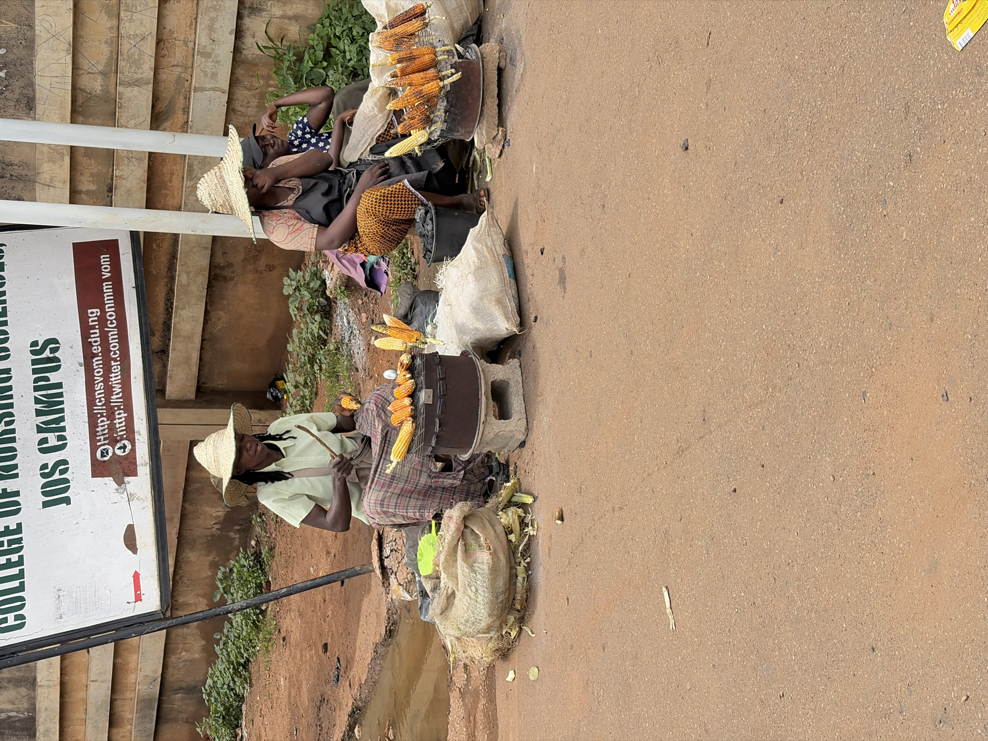 The image shows a street scene in Jos, Nigeria, outside the College of Nursing Sciences, Jos Campus. Two women are selling roasted corn on the side of the road. The woman on the left is wearing a light green shirt, a plaid skirt, and a straw hat. She is tending to a grill with several ears of corn on it. The woman on the right is wearing a patterned dress and a black apron, also with corn being roasted. A third person, partially obscured, sits behind them. In the foreground, there are sacks of corn husks and corn cobs, along with a charcoal bucket. The ground is a mixture of dirt and asphalt. A puddle of water is visible on the left side of the image. A sign for the college is visible in the background.