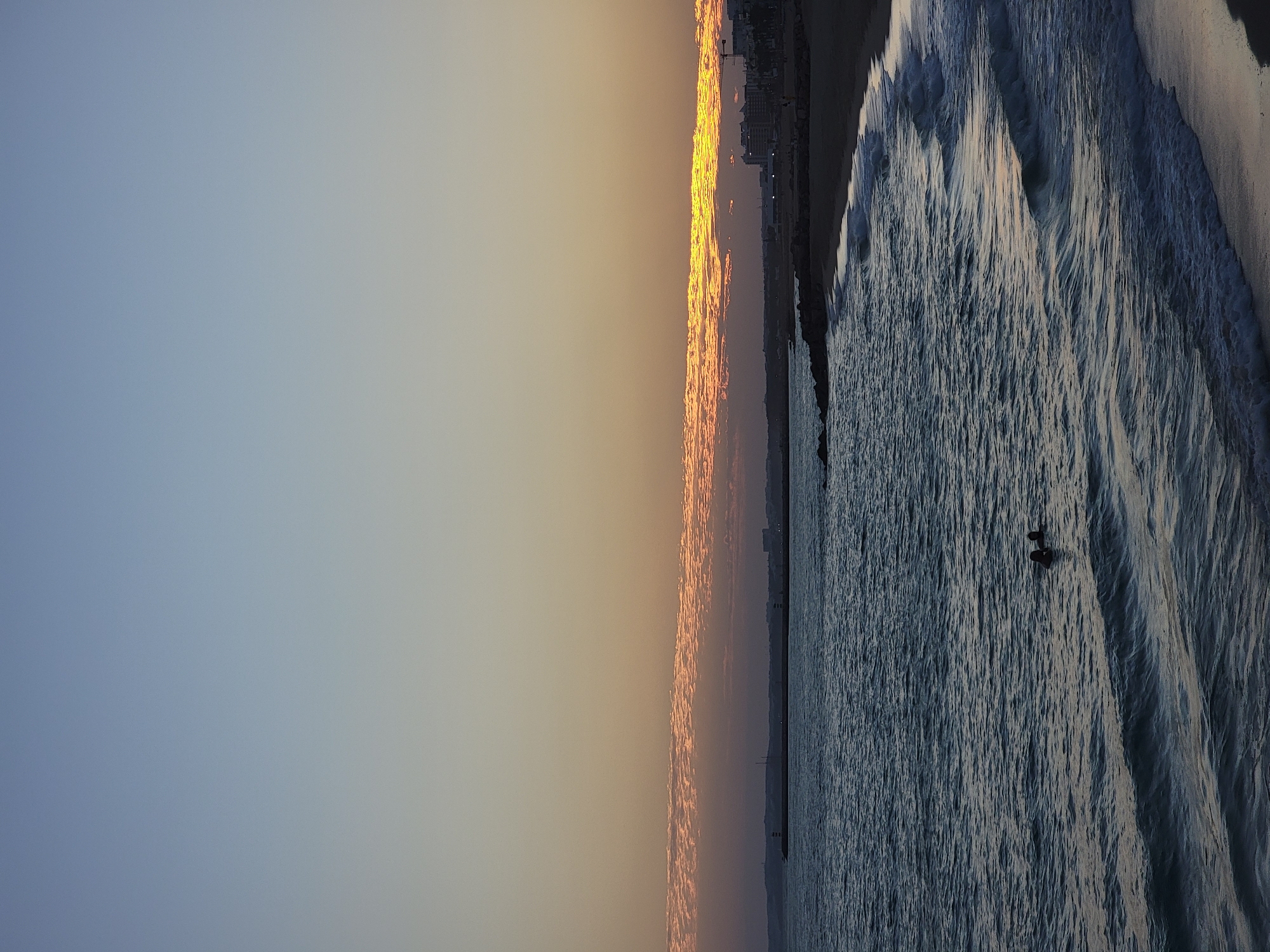 The image depicts a serene sunset scene at the Quarteira beach in Portugal. The sky transitions from a light blue at the top to a warm orange and yellow hue near the horizon, with scattered clouds illuminated by the setting sun. A calm sea with gentle waves rolls onto a sandy beach. In the middle of the ocean, two individuals are visible, appearing to swim or wade in the water. The coastline stretches into the distance, revealing a low-lying urban area with buildings and a pier. The overall atmosphere is tranquil, capturing the end of a day by the sea.