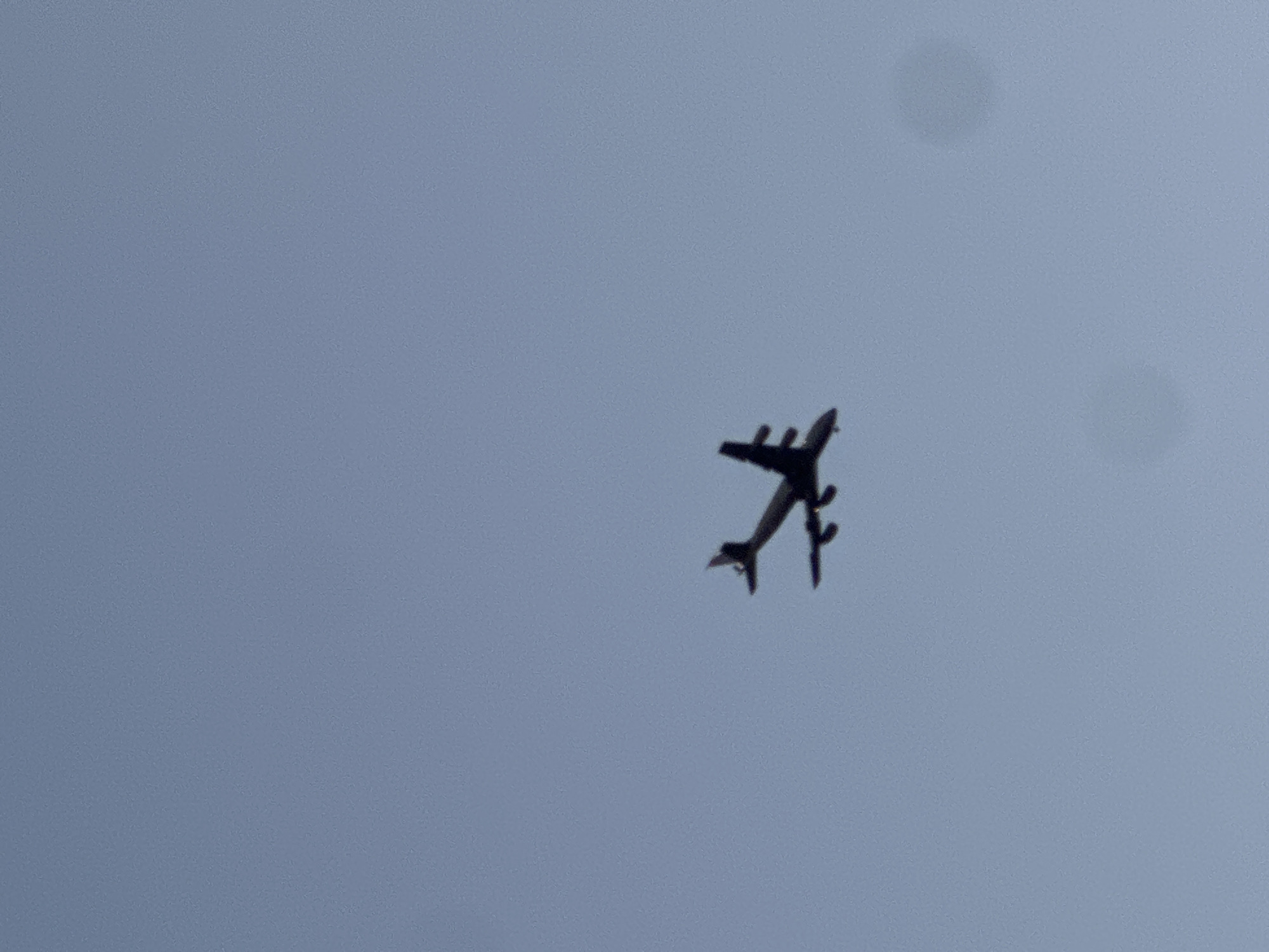 The image displays a dark silhouette of a large, multi-engine airplane flying against a clear, light blue-grey sky. The aircraft is viewed from below, its shape clearly outlined against the lighter background, suggesting it is either very high in the sky or photographed into the light. It appears to have four engines, two under each wing, consistent with a large transport or passenger aircraft. The sky is largely devoid of clouds, showing only a uniform color with some minor graininess and faint, circular bright spots, possibly lens artifacts. The overall scene conveys the impression of an aircraft in flight during daytime. No people or visible text are present in the image. This view was captured over Tel-Aviv, Israel.