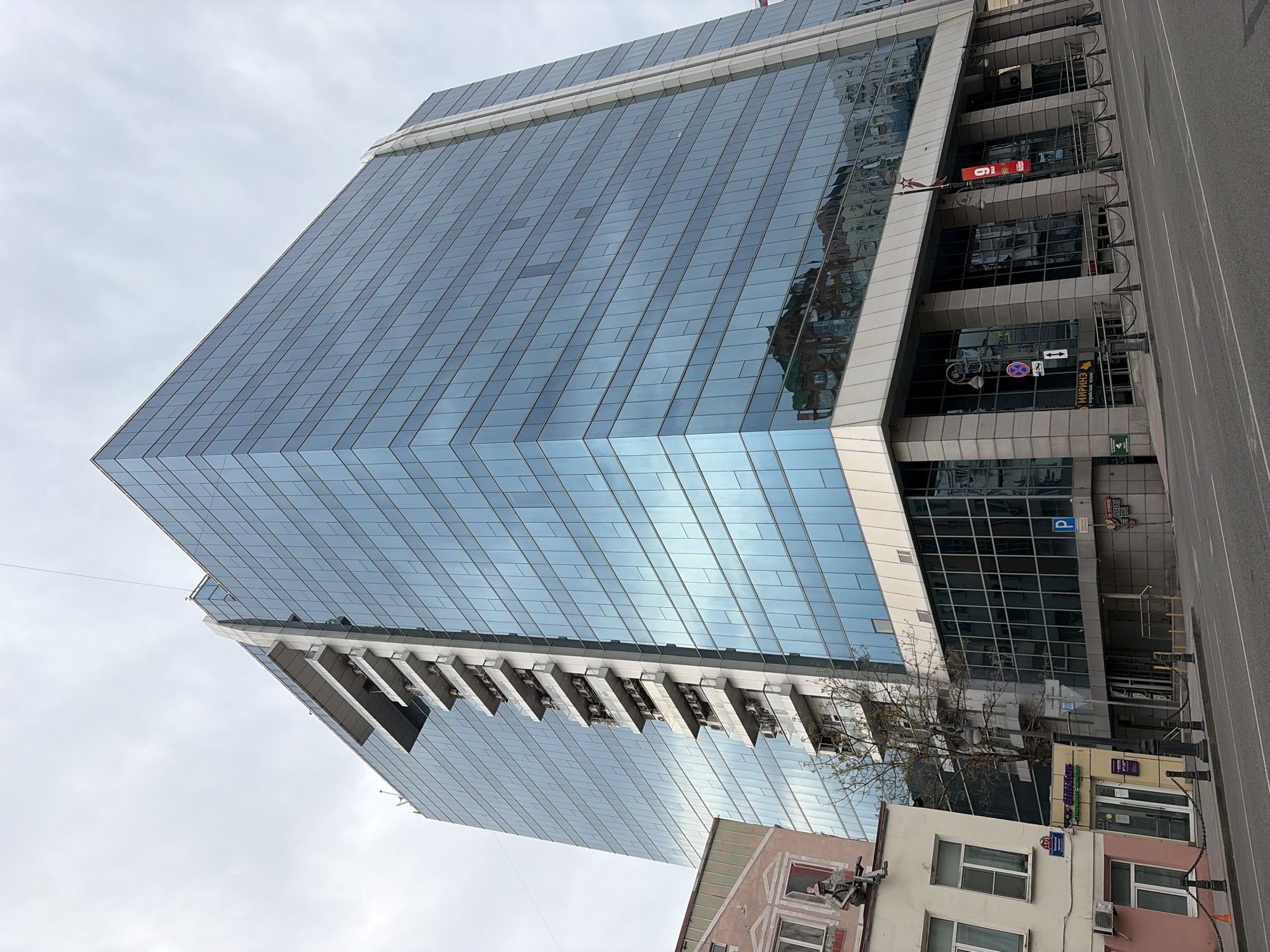 The image captures a tall, modern glass building in Vladivostok, Russia. The building's facade is predominantly glass, reflecting the sky. Below, there is signage in Russian and a parking sign. Street-level retail is visible. A street with barriers is to the right. The angle is from a low vantage point.