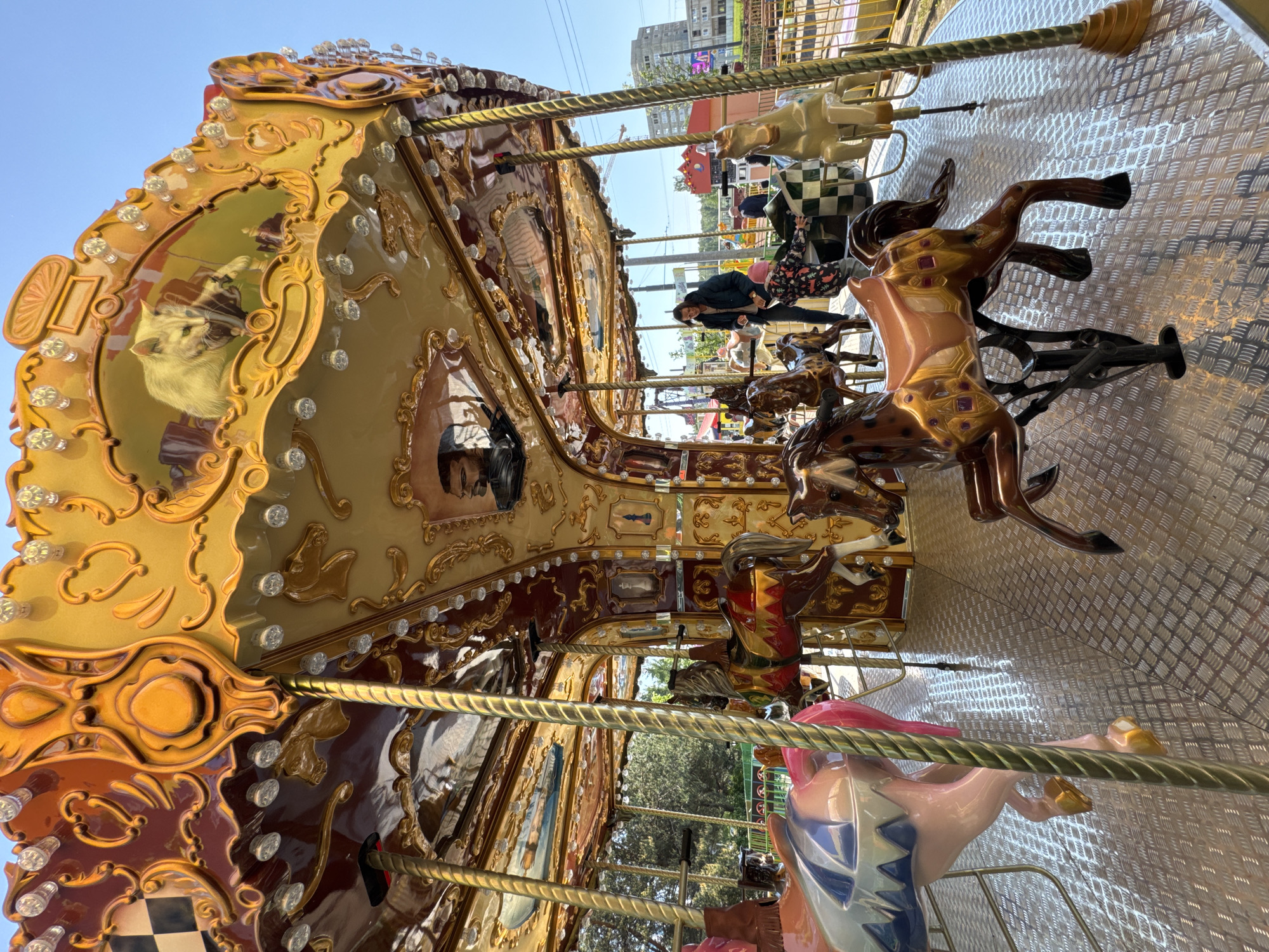 A carousel with golden horses and ornate decorations is in Unknown, Russia. The carousel features animals such as horses, a cat, and people depicted in its painted decorations. The platform beneath the carousel is metal, with diamond-shaped patterns. A woman and a child are riding the carousel. Buildings and trees can be seen in the background. The sky is clear and blue.