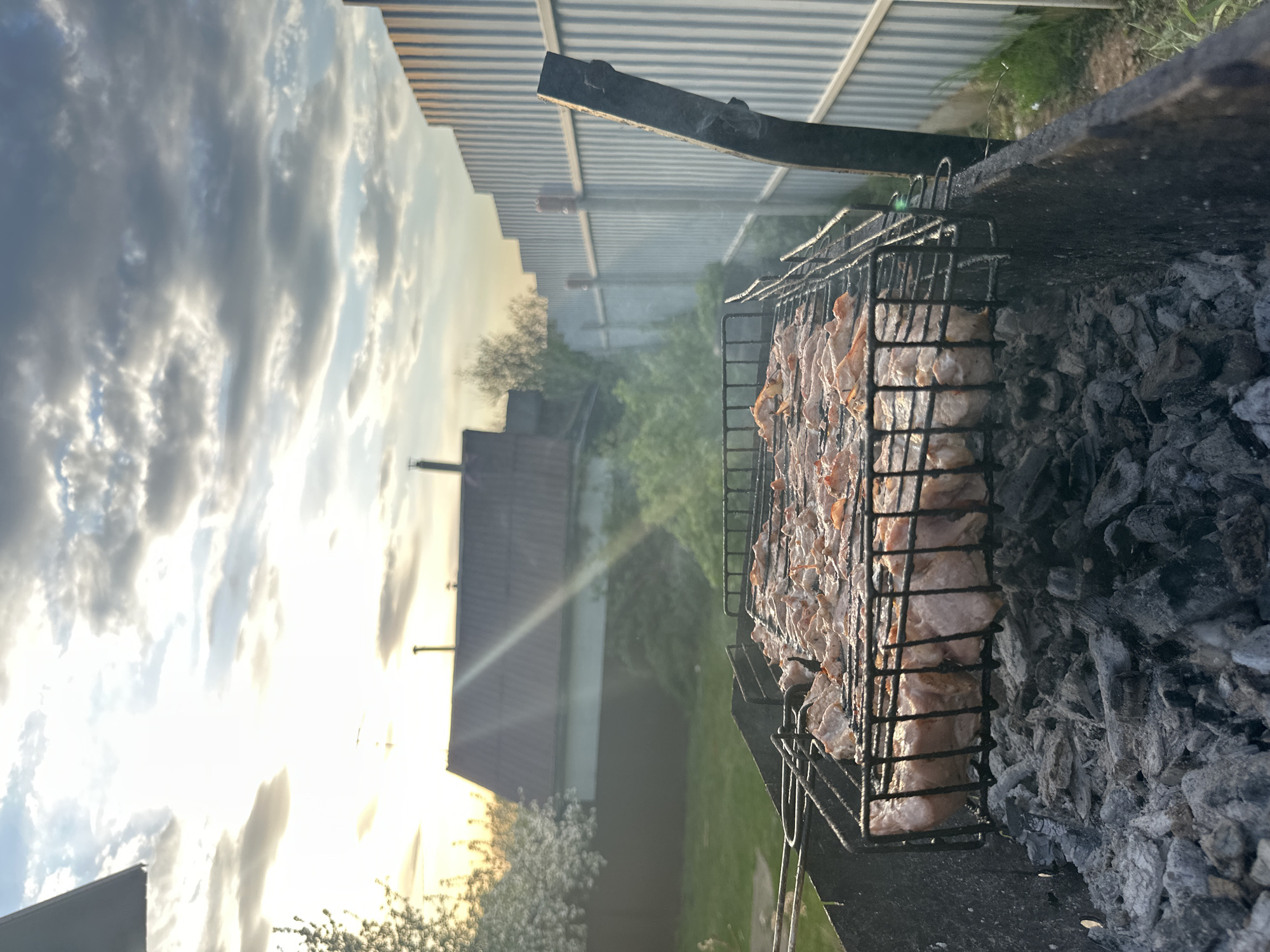 The image depicts a barbecue grill filled with pieces of meat cooking over hot coals. The grill is located outdoors, with a glimpse of a house, trees, a fence, and a cloudy sky in the background. Smoke is rising from the grill. The photo appears to have been taken in the evening, as indicated by the low angle of the sun. It could be in Chaikovsky, Russia.