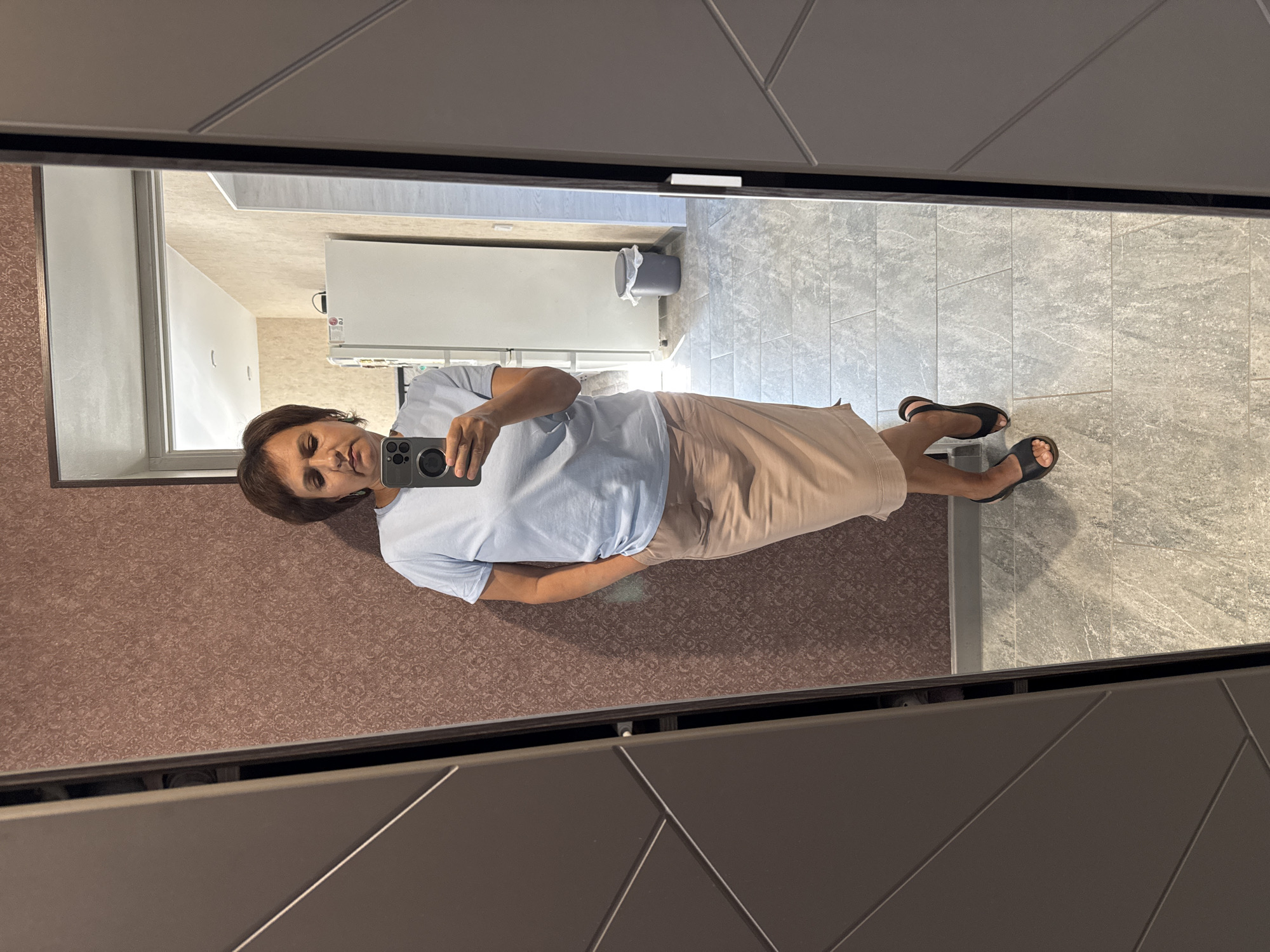 A woman is taking a mirror selfie in a room. She has short brown hair and is wearing a light blue t-shirt and a beige skirt. She is standing with one hand in her pocket and the other holding a smartphone, taking the photo. Her shoes are black open-toed sandals. The room appears to be a hallway or dressing area, with a textured brown wallpaper on the left wall and a tiled floor. In the background, reflected in the mirror, is a kitchen with a white refrigerator and a trash can. The image is taken from a slightly high angle, focusing on the woman and her reflection. The presence of a refrigerator suggests this could be a residential building, potentially in the city of Soloneshnoye, Russia.