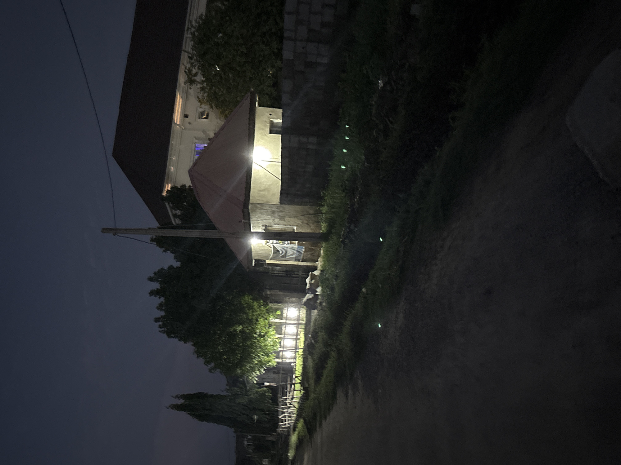 This image depicts a nighttime scene in Angwan Sayawa, Nigeria. An unpaved, dark dirt road occupies the lower left foreground, extending towards the center. To the immediate right of the road, a line of light-colored, possibly wooden, fence posts runs along a patch of illuminated grass and low vegetation. A large, leafy green tree is brightly lit from below, positioned to the left of the central focus. Further left, a tall, slender dark tree stands against the dark blue-grey sky.
In the mid-right of the frame, a small, single-story structure with light-colored walls and a red-brown pitched roof is present. This building is illuminated by two prominent exterior light sources; one lamp is fixed to its front wall, and another is visible under the right-side roof overhang. A blue and white striped fabric is draped on the exterior wall of this small building. A dark utility pole stands directly to the left of this structure, with an overhead wire extending towards the upper right.
Extending from the small building towards the right is a low wall constructed of dark, possibly concrete, blocks. In front of this wall, approximately five small, bright green light points are visible in a linear arrangement within the low vegetation.
In the background, behind the small building and wall, a larger, multi-story building with a dark, expansive pitched roof is discernible. Its walls appear light-colored, and a blue luminescence emanates from a window on its upper floor, indicating an interior light source. Dark, dense foliage surrounds this larger building, blending into the darkened environment. No individuals are visible, and no actions or interactions are depicted. The primary light sources are artificial, illuminating specific areas within the overall dark setting.