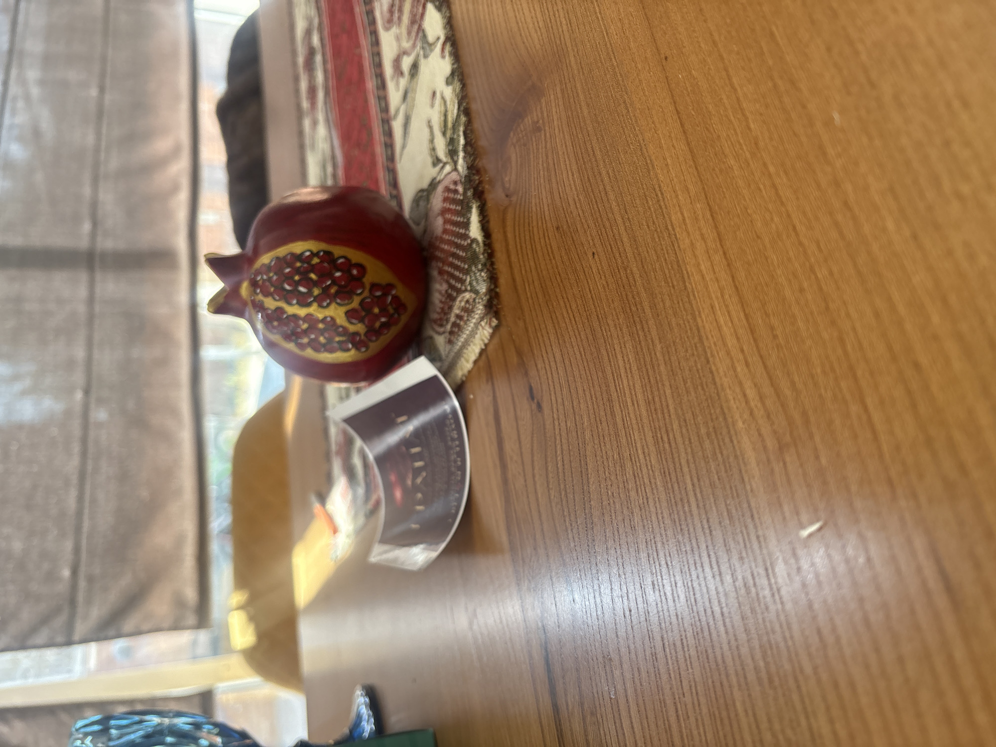 A close-up shot focuses on a table with a wooden surface. On the table, a decorative pomegranate figurine sits on a runner with a red and white floral pattern. The figurine is red with yellow and red detailing on one side, resembling pomegranate seeds. A partially visible tag or label with text is next to the figurine. In the background, a window with blinds is visible, allowing sunlight to stream through and cast a warm glow. The scene appears to be indoors, likely in a home or dining setting. No people are visible in the image.