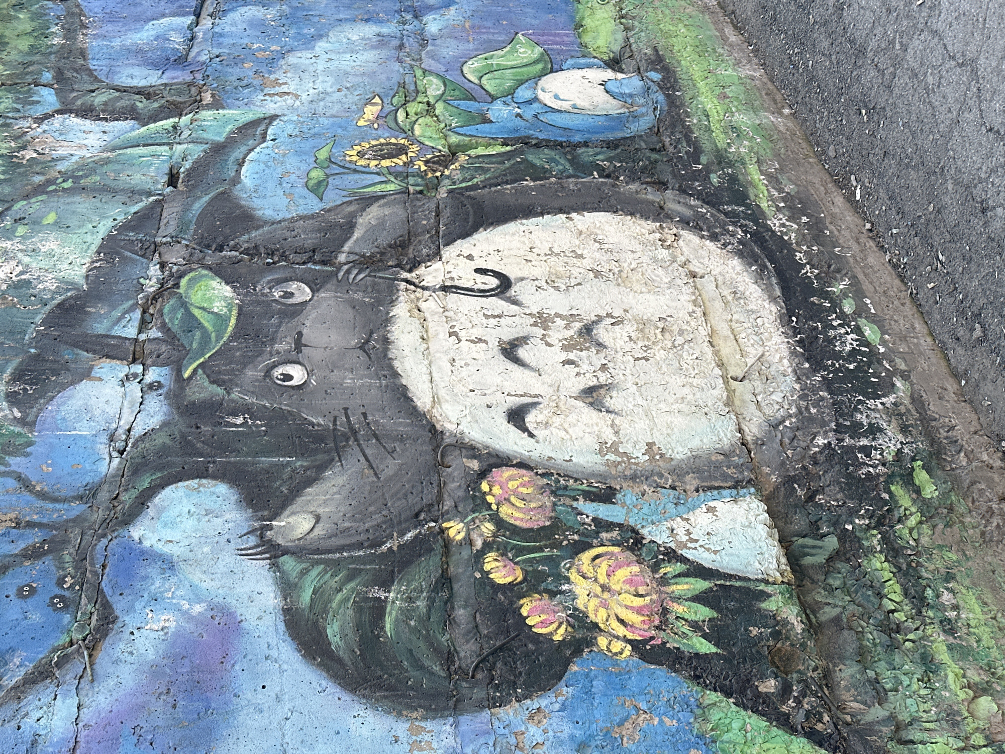 A close-up shot of a concrete slab with a mural painted on it. The mural depicts the character Totoro from the Studio Ghibli film "My Neighbor Totoro". He is grey and has big eyes, as well as a white stomach with small black markings. There are flowers, foliage and blue birds in the mural. The paint is heavily weathered and peeling. The concrete slab is cracked. This mural is located on a building exterior in the city of Novosibirsk, Russia.