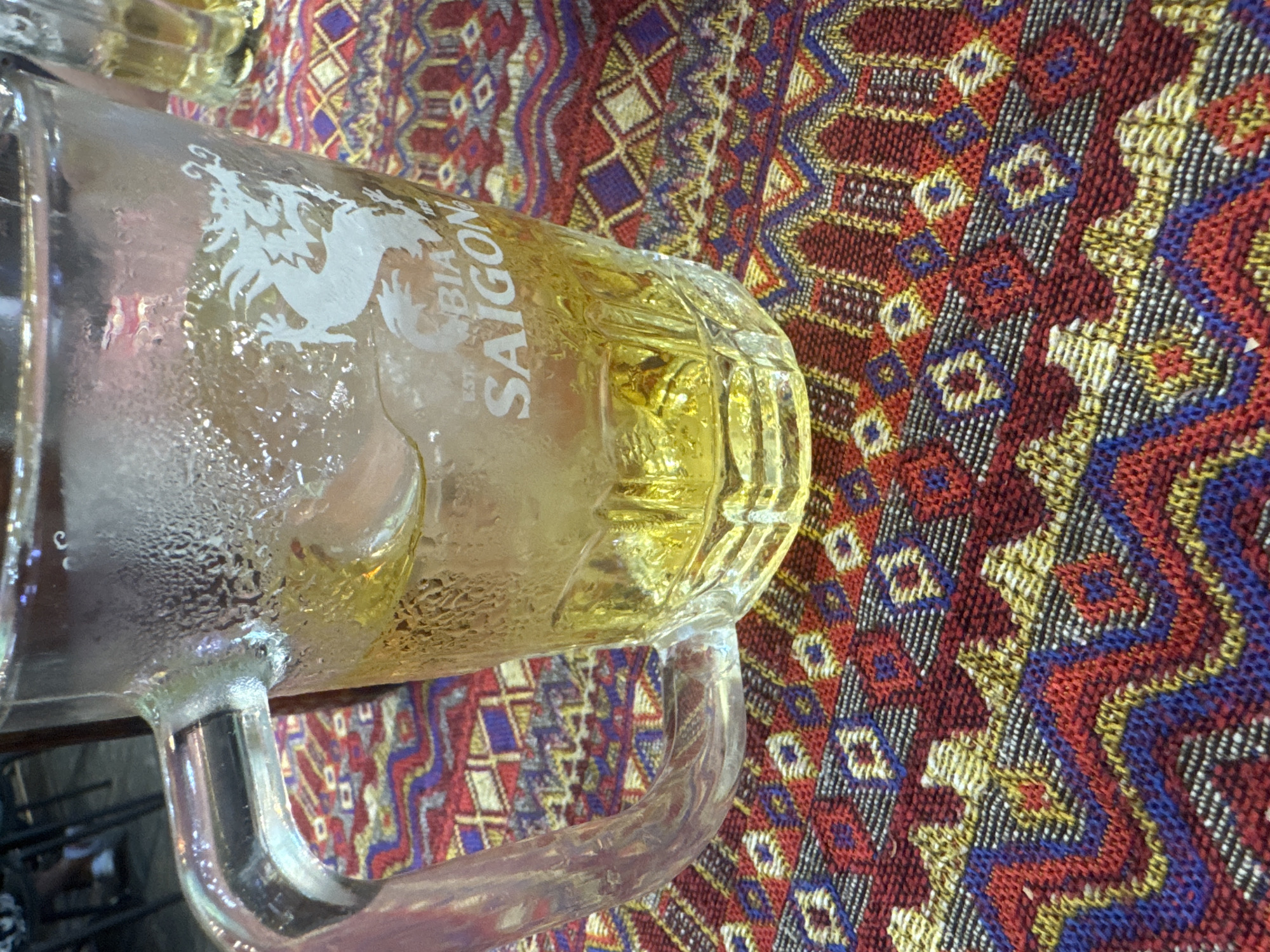 A glass beer mug, labeled "Bia Saigon," rests on a multi-colored patterned tablecloth. The mug appears to be partially filled with a light-colored liquid, likely beer, and shows condensation, indicating it is chilled. The intricate design of the tablecloth includes patterns in red, blue, yellow, and white. The location is Nha Trang Ward, Vietnam.