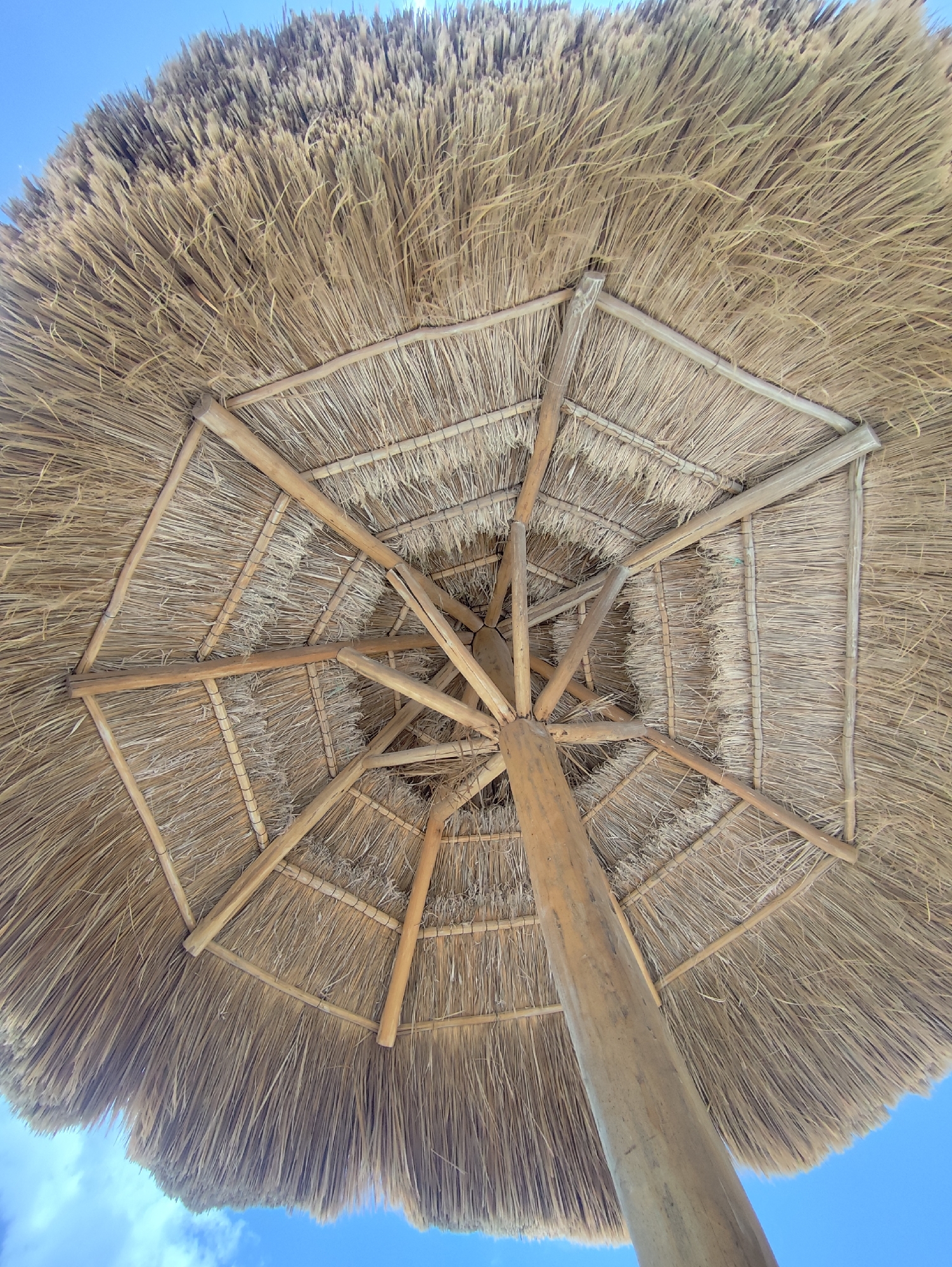 The image, taken from beneath, shows the interior structure of a palapa, a type of open-sided dwelling with a thatched roof made of dried palm leaves. The supporting structure consists of a central wooden pole and radiating wooden beams, arranged in a geometrical pattern of interconnected octagonal frames. The thatching is visible both on the interior and as the overhanging exterior. The sky, pale blue with scattered white clouds, is visible around the edges of the roof. This is a common scene at beaches and resorts in Playa del Carmen, Mexico, where palapas provide shade. No people are visible.