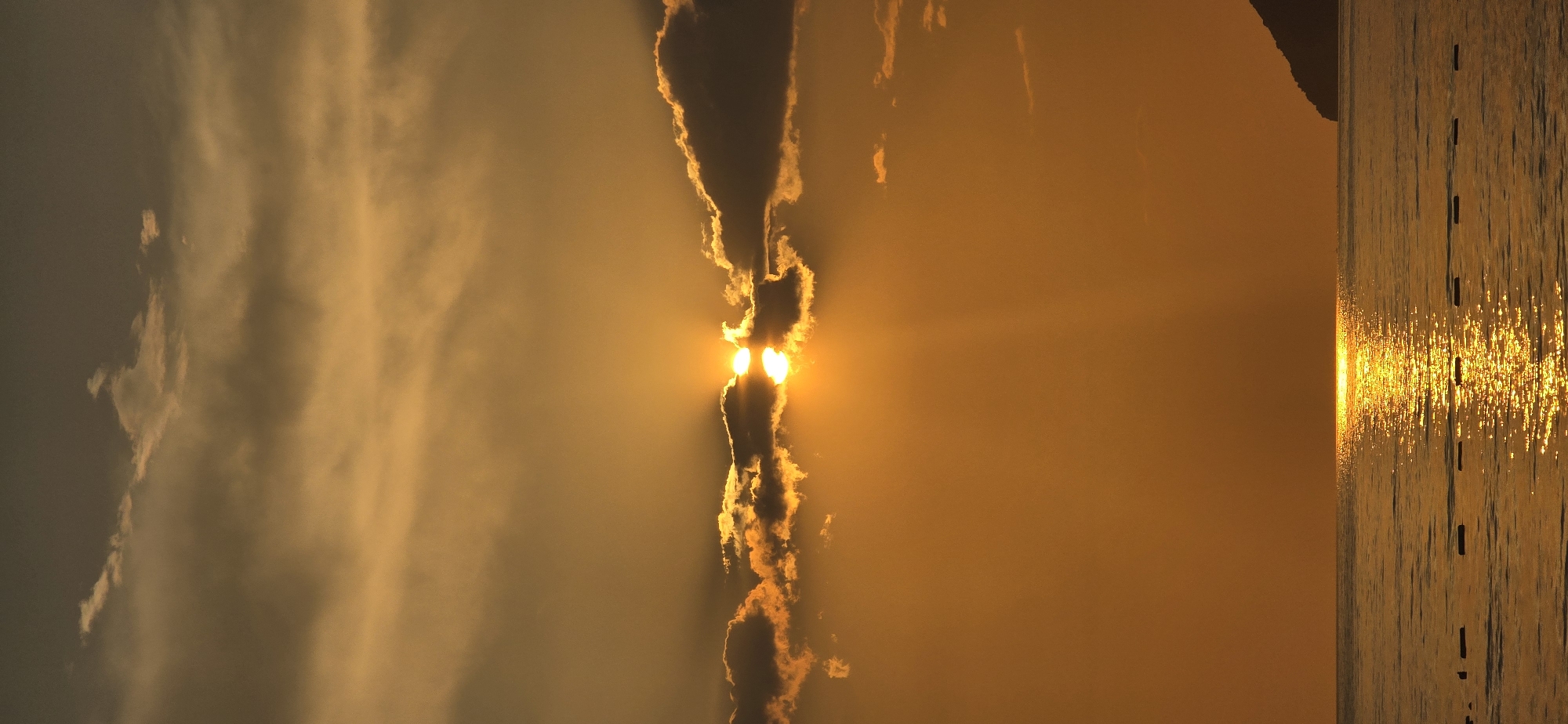 The image captures a golden sunset over the Adriatic Sea in Bar, Montenegro. The sun, partially obscured by clouds, creates a striking visual with its light beams piercing through. The sea reflects the sun's glow, resulting in shimmering highlights across the water's surface. In the water, there are remnants of wooden posts, perhaps from an old pier or structure, standing in a line into the distance. A dark land mass is discernible along the far right edge of the image. The overall scene evokes a sense of serenity and natural beauty.