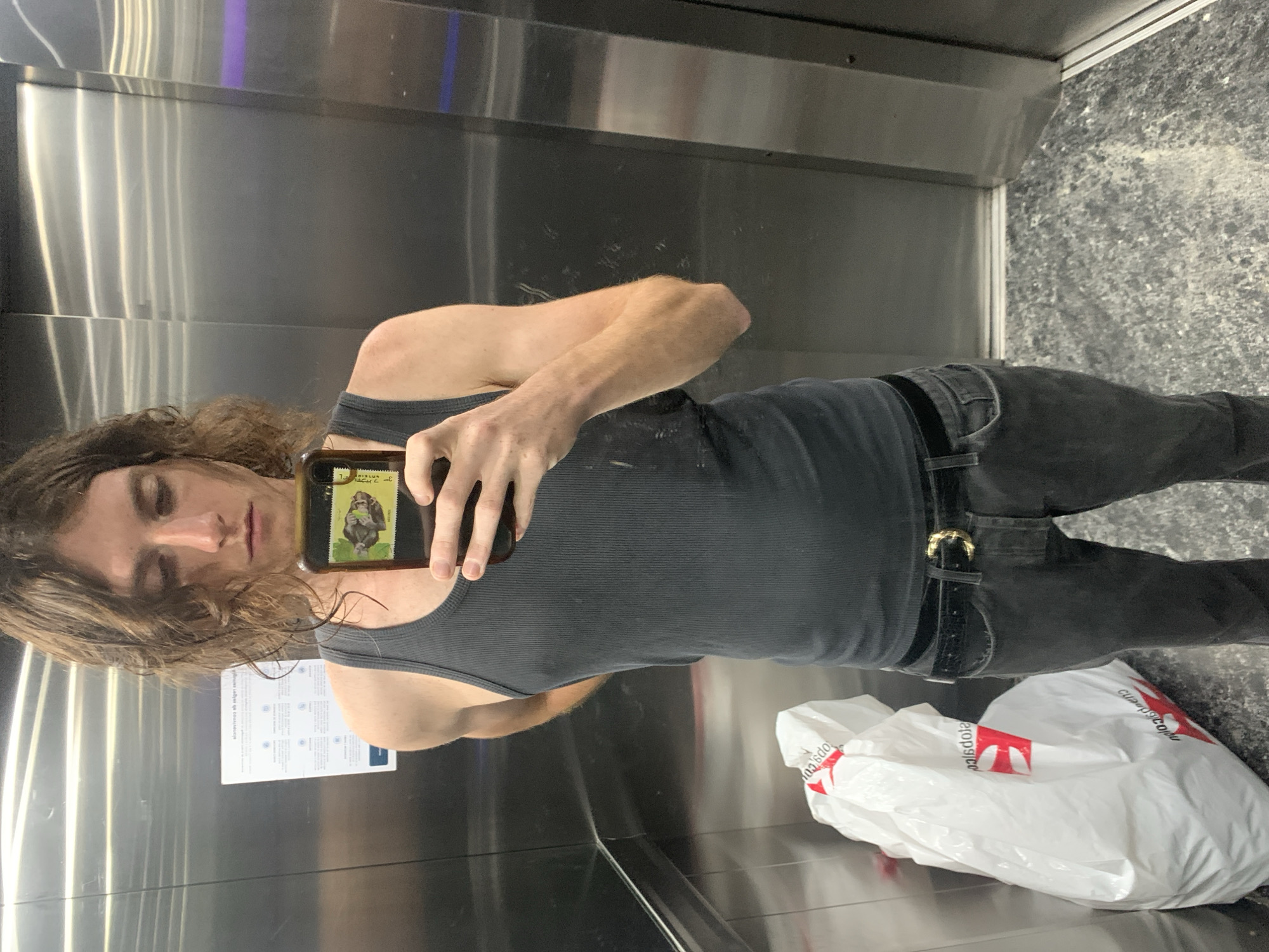 A person is taking a selfie in an elevator. The person has long brown hair and is wearing a grey tank top and black jeans with a black belt and gold buckle. They are holding a phone with a stamp of a monkey on the back. A white plastic bag with red markings, possibly from a pharmacy called "Farmacity," is on the floor next to them. The interior of the elevator is stainless steel with a marble floor, placing the location likely in the city of Buenos Aires, Argentina due to the reference to the local pharmacy.