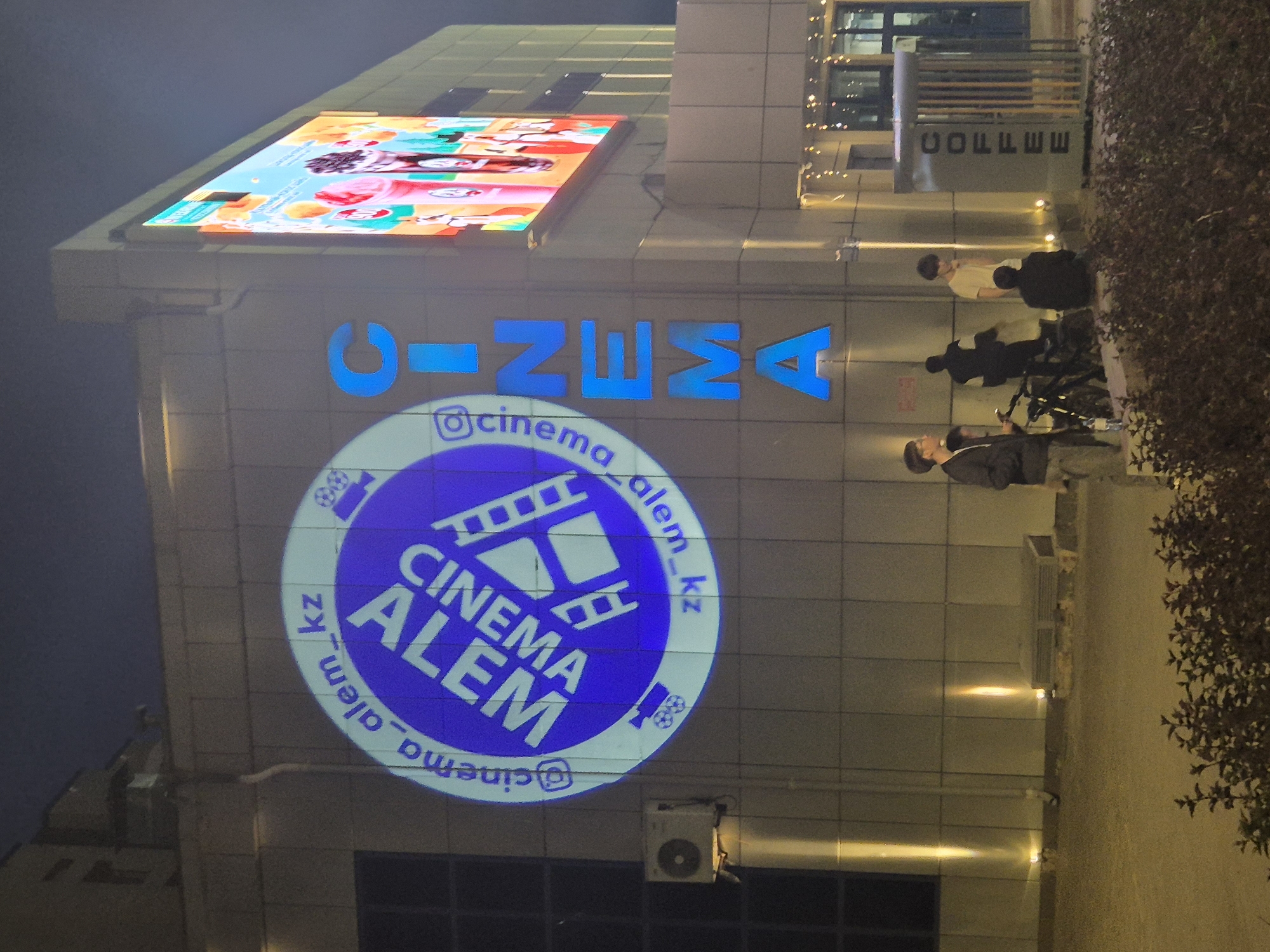 The image depicts the facade of the Cinema Alem building in Kokshetau, Kazakhstan. Above the entrance, in blue block letters, is the name of the cinema "CINEMA ALEM." Below this, a circular sign illuminated on the building reads "@cinema_alem_kz," encompassing a film reel graphic and the words "CINEMA ALEM." The building has a large digital display above the cinema's name, showing animated content.
To the right of the building entrance, three individuals are present. Two stand near parked bicycles, while a third is slightly further away. Above them, "COFFEE" is written vertically on the building. An air conditioning unit is visible on the facade near the cinema logo. The building's exterior is clad in light-colored panels. The adjacent area is marked by a bush or tree line, and the overall lighting suggests it is dusk or evening.