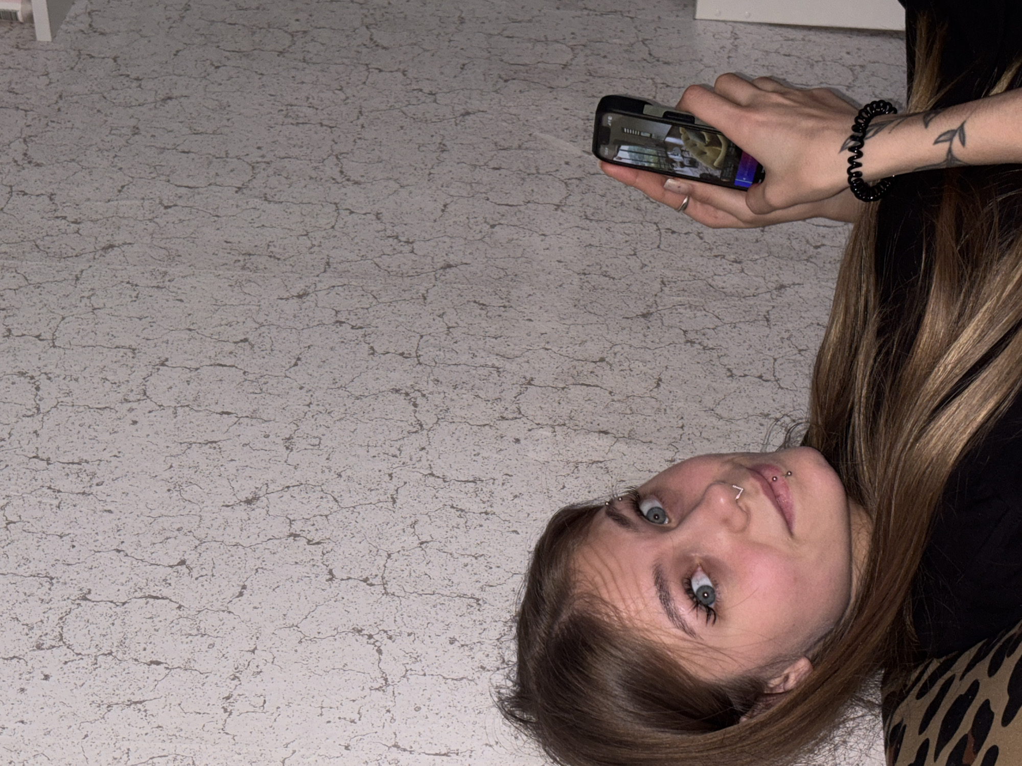 A young Caucasian female is lying on a light-colored, cracked surface, possibly tile or linoleum, while holding a black smartphone in her right hand. The phone screen displays an image. The female has fair skin, light brown hair with blonde highlights, blue eyes, and piercings on her eyebrow, nose, and lip. She is wearing a black shirt and a black spiral hair tie on her right wrist, which also has a tattoo of leaves. Part of a leopard-print pattern is visible on the bottom right of the frame. The location is not discernible, and there is no information to identify the scene as being in Фоки, Russia.