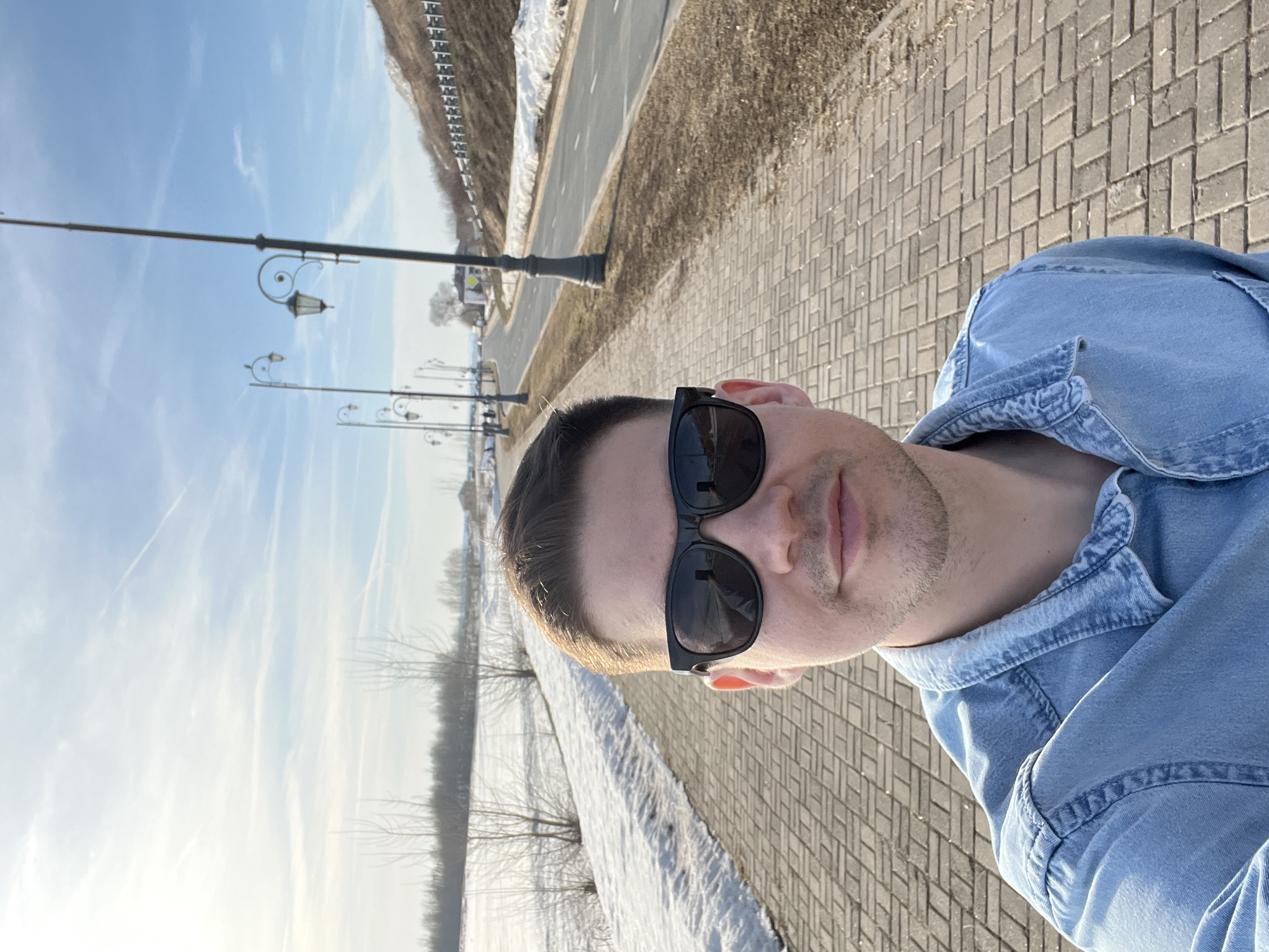 The image depicts an outdoor selfie taken in Sviyazhsk, Russia, featuring a male subject in the foreground. The man, appearing to be in his late 20s to early 30s, has short brown hair and is wearing black rectangular sunglasses and a light blue denim shirt. He is positioned center-frame, looking directly at the camera with a subtle smile.
He stands on a light-colored brick-paved path, laid in a herringbone pattern. To the left of the path, a snow-covered field with patches of visible brown earth extends into the distance, punctuated by numerous bare deciduous trees and shrubs. A dense line of trees forms the horizon on this side. To the right of the path, an asphalt road with a dashed white centerline runs parallel to the path. A series of dark, ornate streetlights, featuring double arms with lantern-style lamps, are regularly spaced along the roadside. Beyond the road, an upward-sloping embankment, partially covered in snow and dry grass, is visible, topped by a metal guardrail. The sky is clear blue with sparse, wispy white clouds and faint contrails. The scene is brightly illuminated by sunlight, suggesting late winter or early spring conditions.