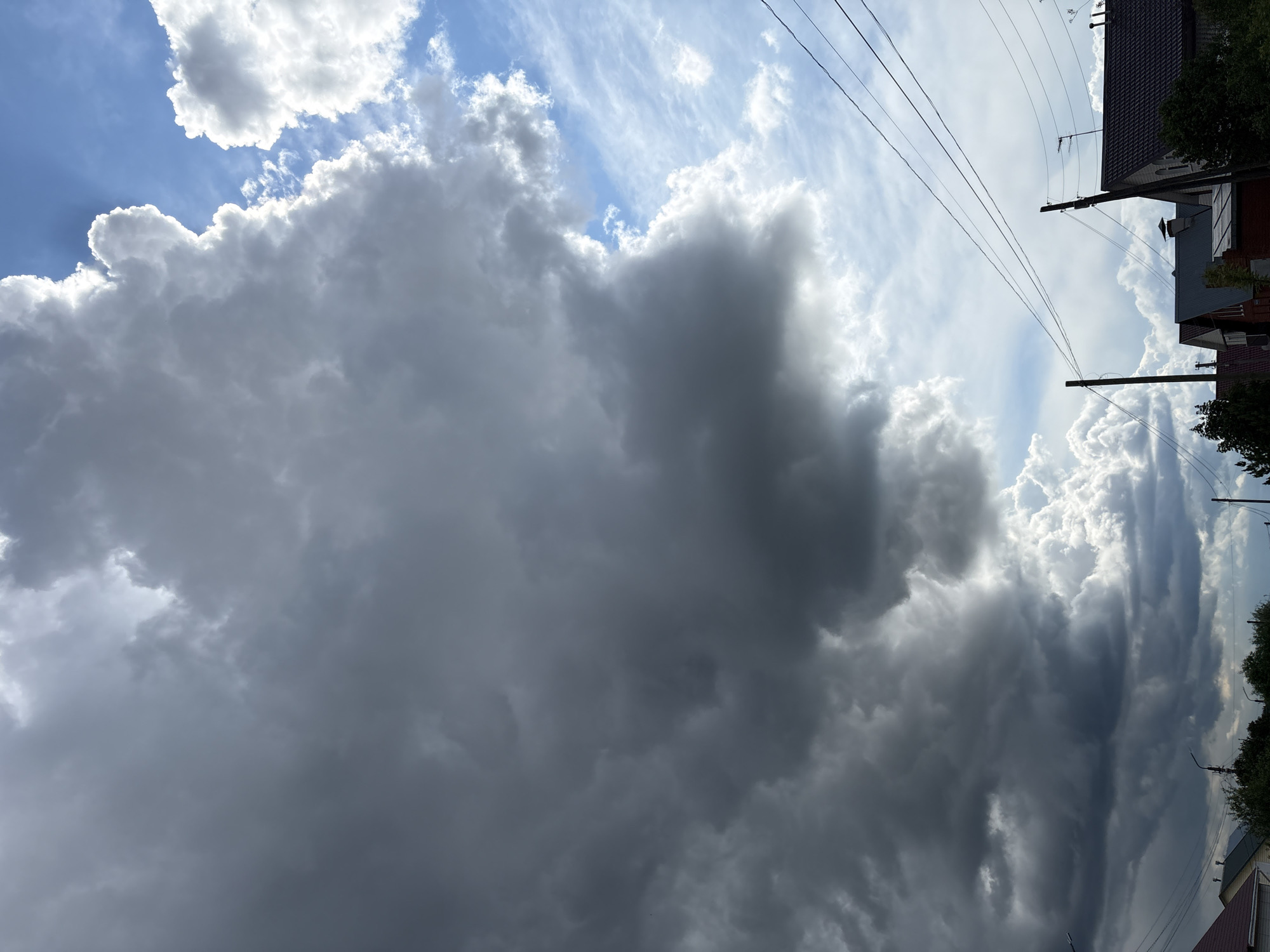 The image shows a view towards the sky, dominated by large, dense storm clouds. Part of a residential area in Прикамский, Russia is visible in the upper right corner, including rooftops and power lines. The sky is partially visible, with white and blue hues visible between the clouds. The image seems to be taken from a low angle, looking upwards.