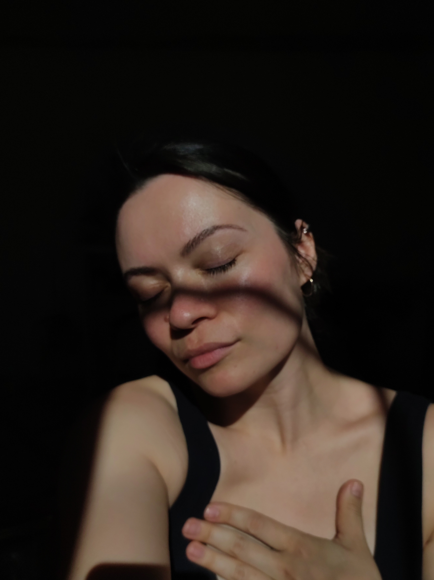 A woman with dark hair is in a dimly lit area. She is wearing a black tank top. Her eyes are closed, and there is a shadow across her face. She has earrings in her left ear, and her right hand is resting on her chest. The background is dark. There is no clear indication of the location being Фоки, Russia in the image.