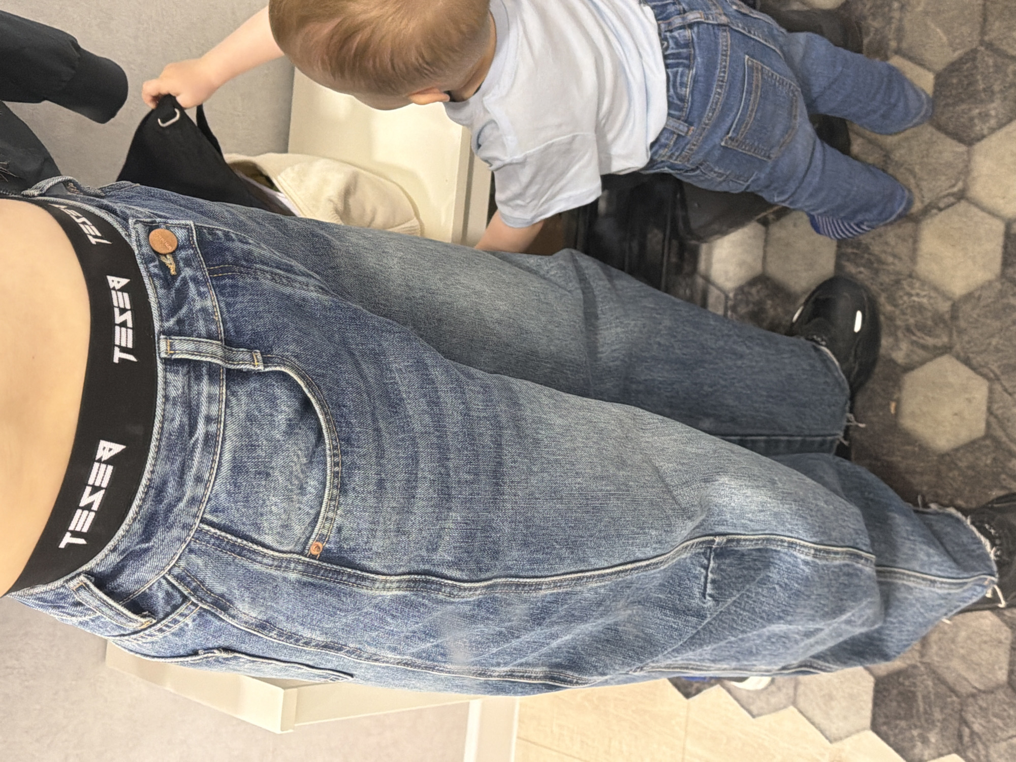 The image shows a partial view from above of a person wearing blue jeans and black shoes. The waistband of the jeans is visible, revealing a black band with the word "DIESEL" printed in white. A young child, wearing a blue t-shirt and blue jeans, is partly visible interacting with a black bag on the person's lap. The background includes a white surface, potentially a piece of furniture, and a tiled floor with a hexagonal pattern. The location and connection to the city of Votkinsk, Russia, cannot be determined from the visual information provided.