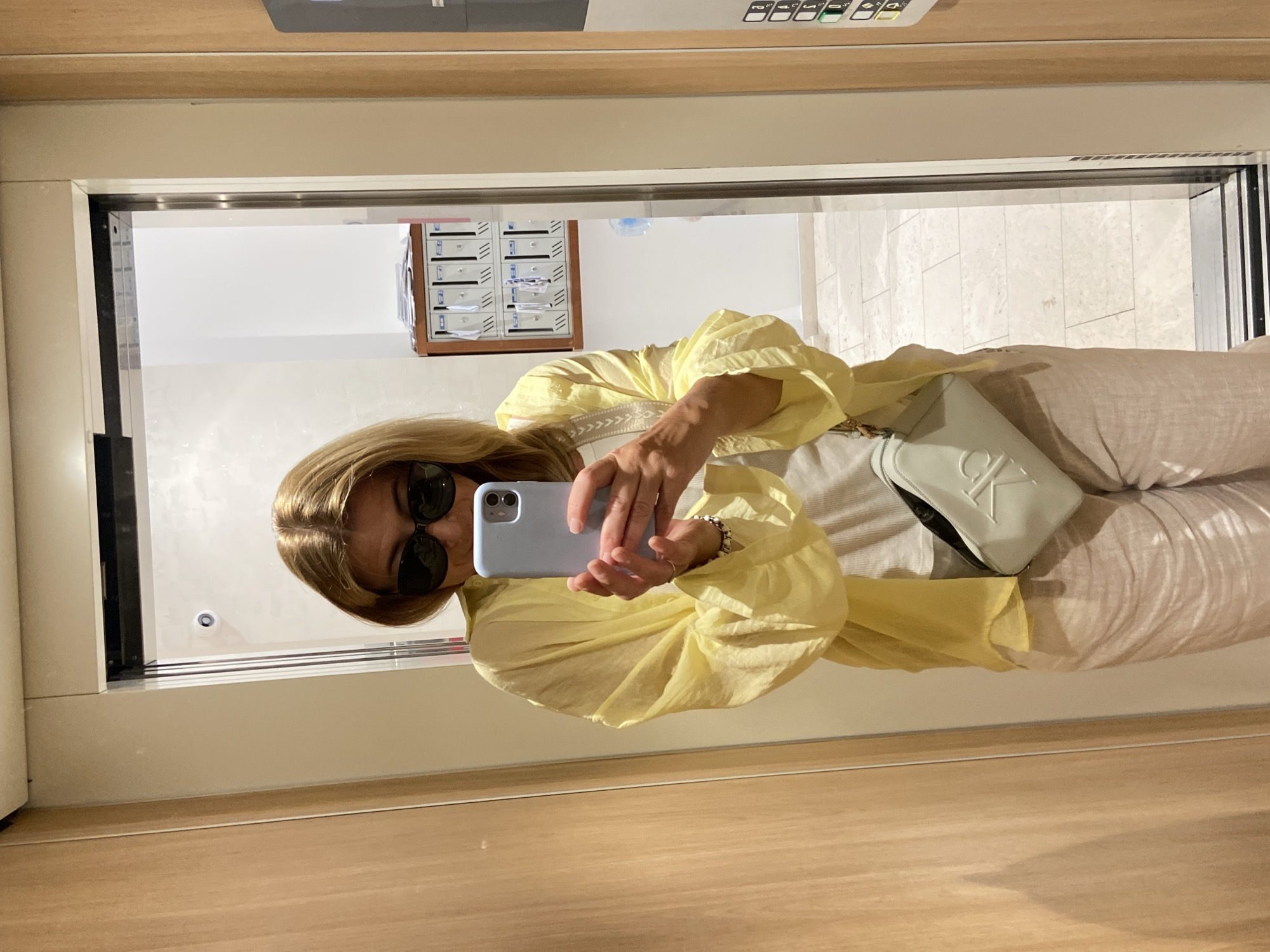 A person is taking a selfie in a mirrored space. The reflection shows a light-skinned person with blonde hair wearing sunglasses and a yellow top. The person is holding a light blue smartphone. The background visible through the mirror includes a mailbox system and a white tiled floor.
While the image does not explicitly state the location, the presence of European-style mailboxes and general architectural styles suggest the photo could be taken in an apartment complex in Budva, Montenegro or similar European locales. There is no conclusive evidence, however, to confirm this.