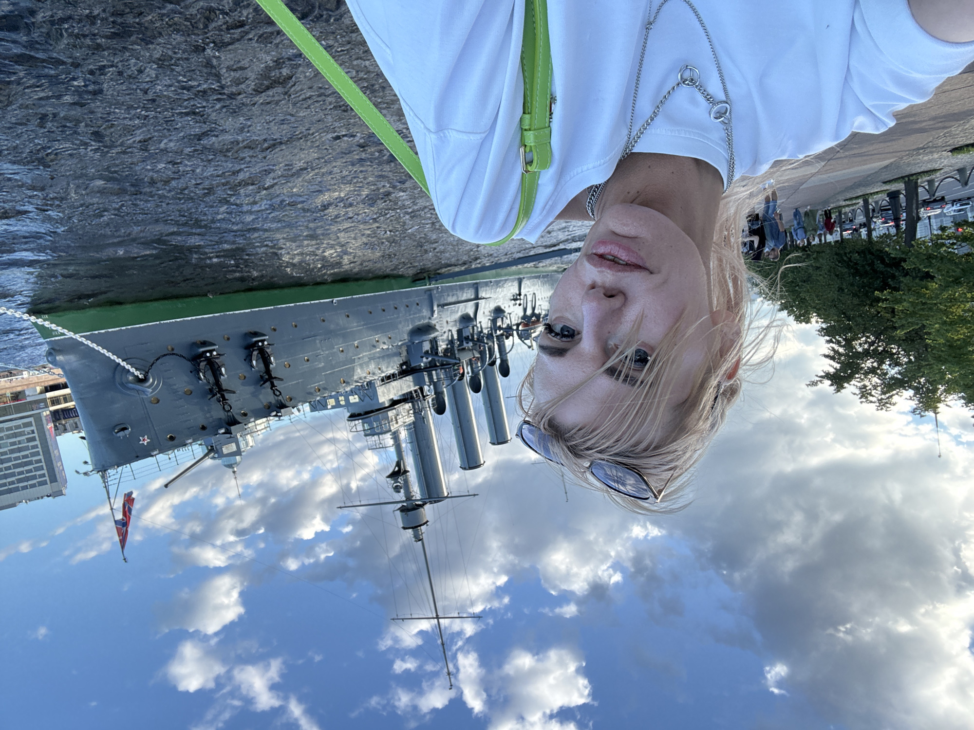 The image shows a woman taking a selfie in front of the cruiser Aurora in Saint Petersburg, Russia. The cruiser, a historic Russian warship, is docked on the Neva River. The woman is wearing a white t-shirt and a silver necklace. She has blonde hair and is wearing sunglasses pushed up on her head. In the background, there is a sidewalk with people walking, trees, and buildings. The sky is cloudy with patches of blue. The water of the Neva River is visible in the foreground.