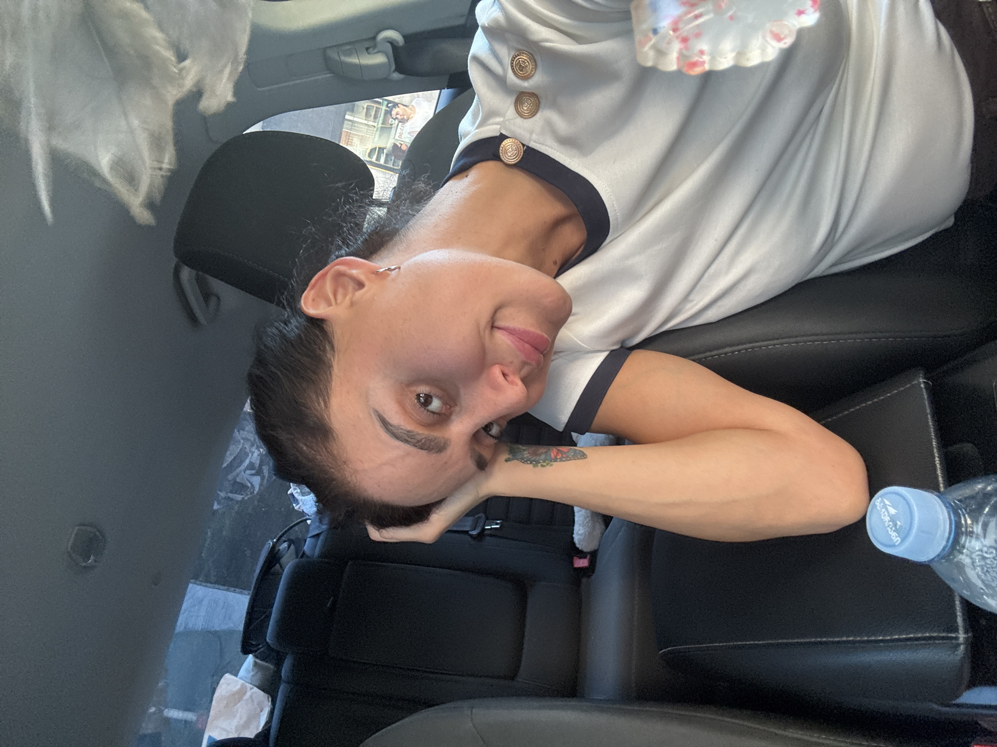 In a selfie taken inside a car, a woman with dark hair is positioned in the passenger seat. She is looking towards the camera, with her left arm resting on her head and her hand near her forehead. Her eyes are open, and she has a slight smile. She is wearing a white t-shirt with a navy collar and gold buttons. A tattoo of a butterfly can be seen on her left forearm. A bottle of water with a blue cap is visible in the center console. The background shows the car's interior, including the back seats and the ceiling. The car appears to be parked, possibly in the city of Tbilisi, Georgia, given the visible architecture and people outside the car window.