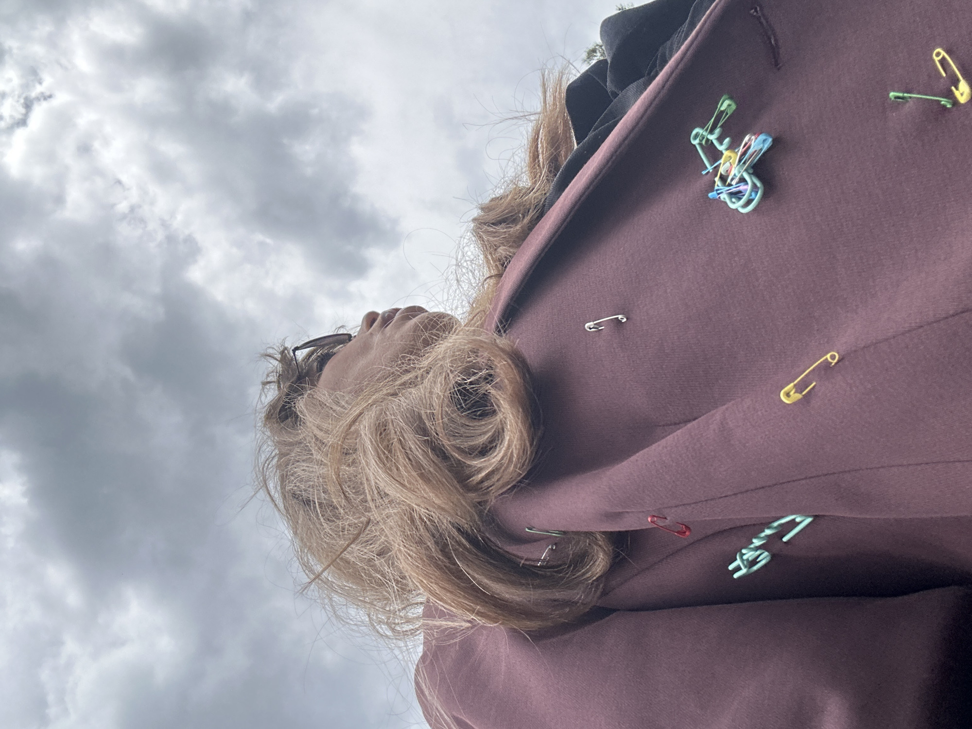The image, taken from a low-angle perspective looking upwards, features a person partially visible against an overcast sky, likely in Tbilisi, Georgia. The main subject is the upper body of an individual, seen primarily from the side and back. Their head is tilted upwards, gazing towards the sky, with portions of their profile, including the nose and chin, and glasses frames visible. The person has light blonde or light brown hair, which appears curly or wavy, cascading over their shoulders and back.
They are wearing a dark reddish-brown or plum-colored jacket or coat. A distinctive feature of the garment is the array of safety pins attached to it. These pins vary in color, including standard metallic silver, bright yellow, green, light blue, and red. A prominent cluster of multiple light blue, yellow, and red safety pins is arranged intricately on the upper left side of the jacket, resembling a decorative element. Other colored pins are scattered individually across the fabric.
The background consists entirely of a cloudy, grey sky, indicating an overcast or partly cloudy daytime setting. No direct sunlight is visible. There are no specific architectural or landscape cues within the image itself to identify the location as Tbilisi, Georgia, beyond the provided context. No text is visible in the image.