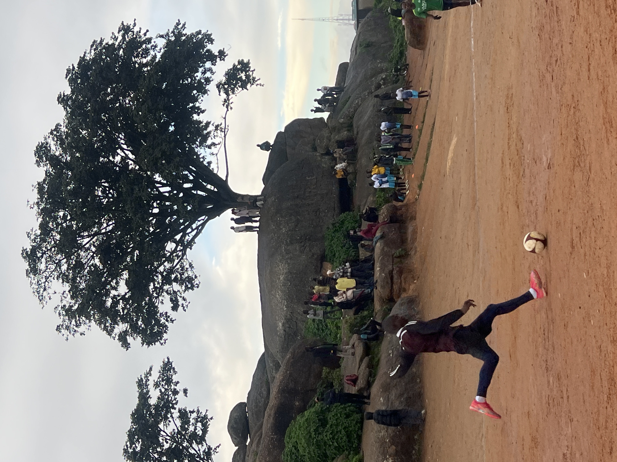 The image depicts a group of people gathered at a rocky outcrop with a large tree overhead, likely in Jos, Nigeria. In the foreground, a young Black male, wearing a maroon jacket with white stripes on the sleeves and black pants, is in mid-stride, kicking a white and brown soccer ball. He is wearing bright pink athletic shoes. The soccer ball is positioned to the right of his kicking foot. A second soccer ball is visible further in the foreground. The ground is a reddish-brown, dusty dirt surface, suggesting a playing field.
In the background, a significant number of people are visible, some standing and others sitting on the rocks. They appear to be observing the soccer activity. The rocks are large and uneven, covered in patches of green vegetation. A large, mature tree with a wide canopy dominates the upper portion of the scene, casting shadows. The sky is overcast with some lighter patches. Further in the distance, a cell tower or radio mast is visible on the right side of the frame. The overall scene suggests a casual outdoor gathering and a soccer game taking place in a natural, somewhat rugged environment.