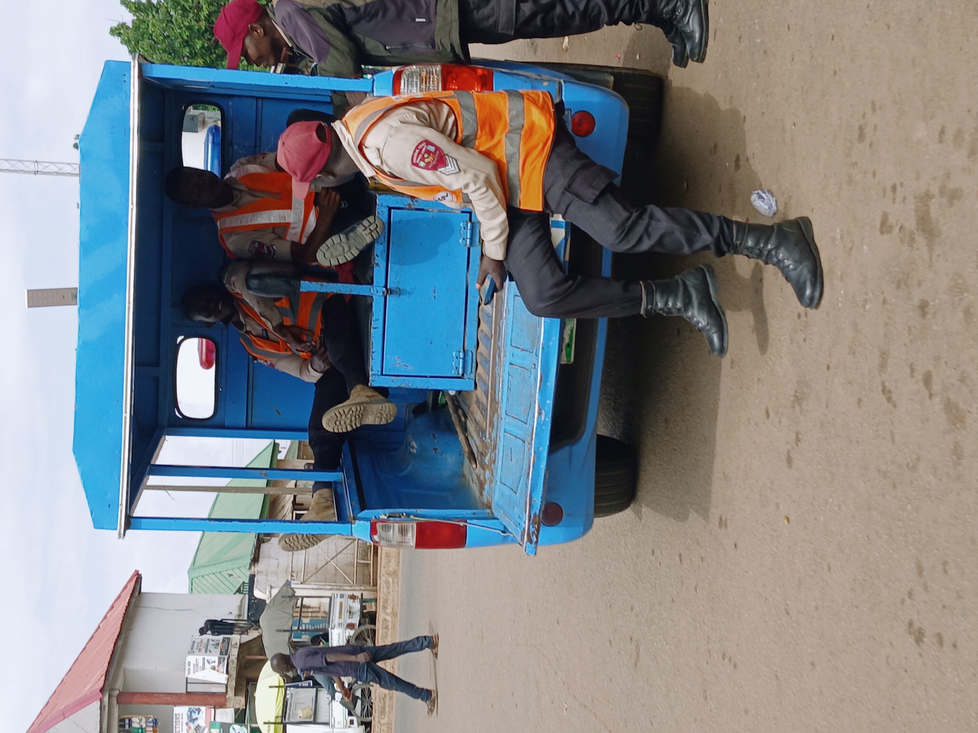 The image shows a blue vehicle with several people in the back. Some of the individuals are wearing orange vests and red hats. The vehicle is on a paved road in what appears to be a city environment, with buildings and a person walking nearby. The specific city is unknown.
