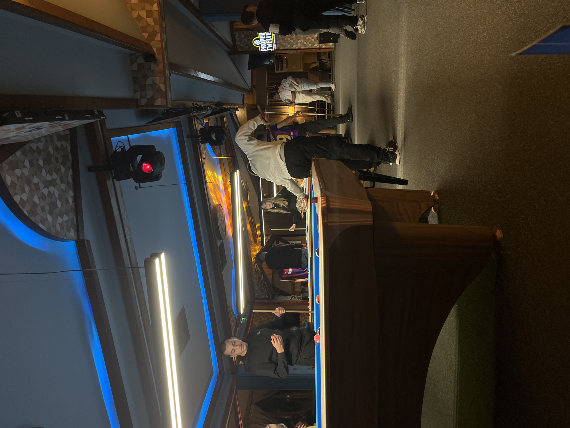 The image shows an indoor scene at the Corner Spot bar in Plovdiv, Bulgaria. Several people are gathered around a billiard table, with some playing pool. One person is in the process of taking a shot, while others are observing. The interior of the bar features decorative lighting, including blue neon accents and patterned wall coverings. There is also a "Proper Twelve" branded sign visible in the background.