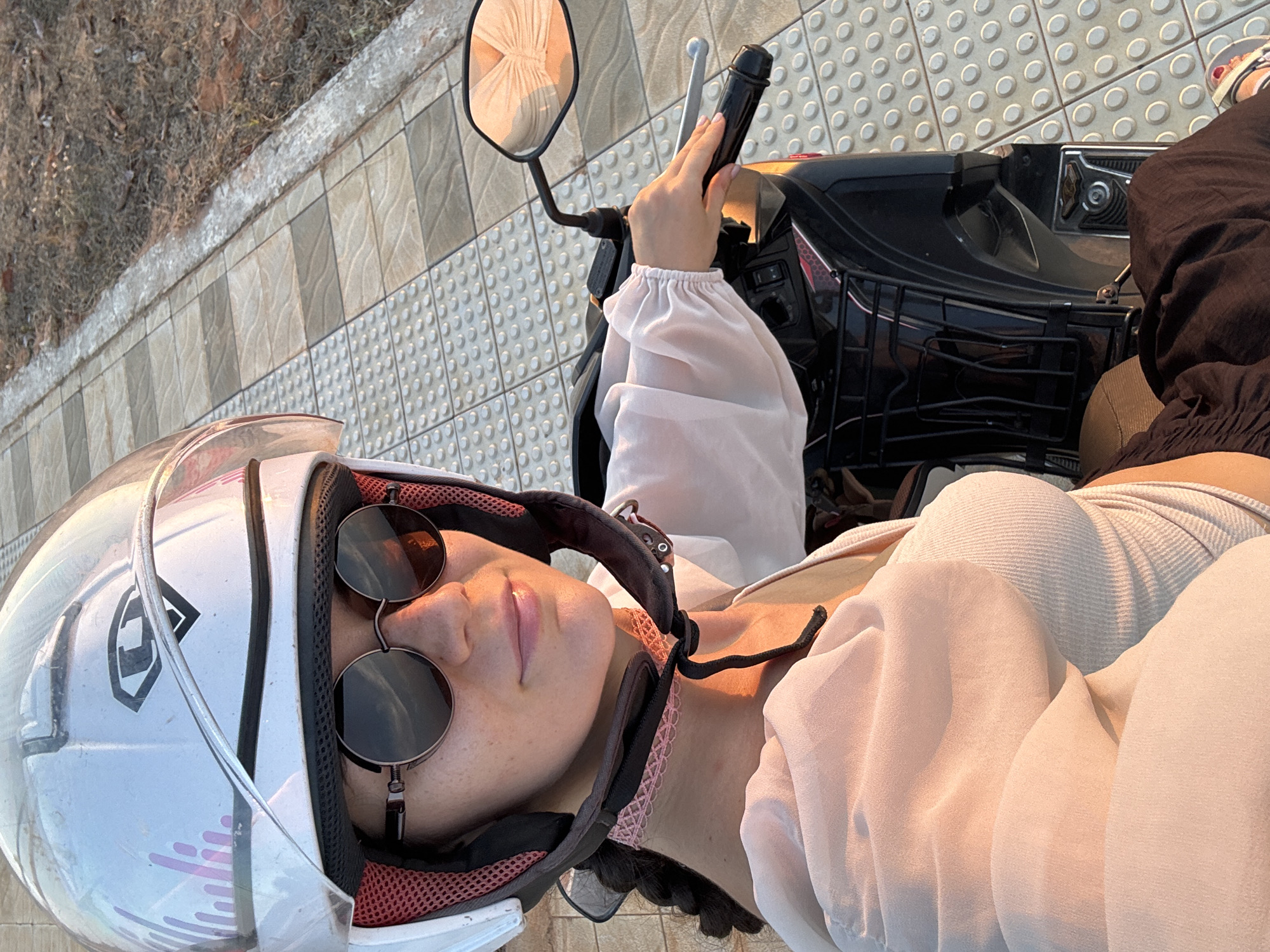 An overhead photograph captures a young woman seated on a black scooter, bathed in the warm light of golden hour, likely in Mũi Né, Vietnam. The scene is set on a tiled sidewalk next to a dry, grassy embankment.
The woman is wearing a white full-face helmet with a clear visor, featuring a black and white geometric logo. She also wears round, dark-rimmed sunglasses over her eyes, with her face gently upturned, lips slightly parted in a relaxed expression. She is dressed in a light-colored, possibly pale pink or white, flowing blouse with puffy sleeves, layered over a ribbed tank top. Her right hand rests casually on the scooter's handlebar, near the brake lever, and a rearview mirror reflects a soft, indistinct fabric. Her legs are clad in loose-fitting dark pants.
The scooter is black, with visible chrome elements and a black wire basket at the front. It is parked on a pavement composed of alternating grey tiles; some feature a subtle wavy pattern, while others are tactile paving with raised circular bumps. The soft, warm light illuminates the scene, suggesting either sunrise or sunset, creating a peaceful and serene atmosphere. No discernible text is clearly visible apart from the helmet logo.
