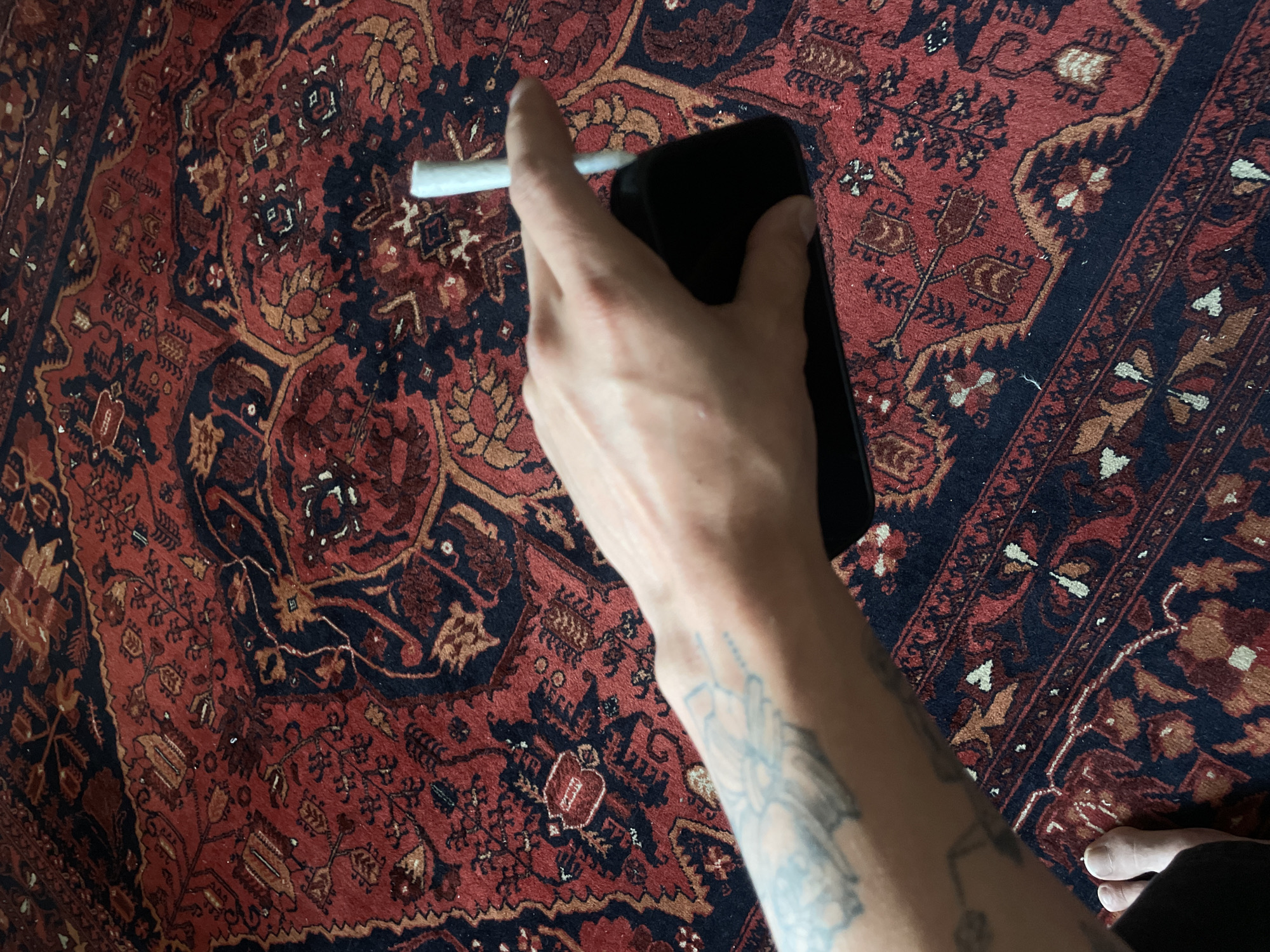 A photograph shows a hand holding a lit cigarette and a smartphone over a rug. The rug is patterned with red, black, and brown colors. The hand has visible tattoos. The background indicates the presence of flooring similar to that of Mexico City.