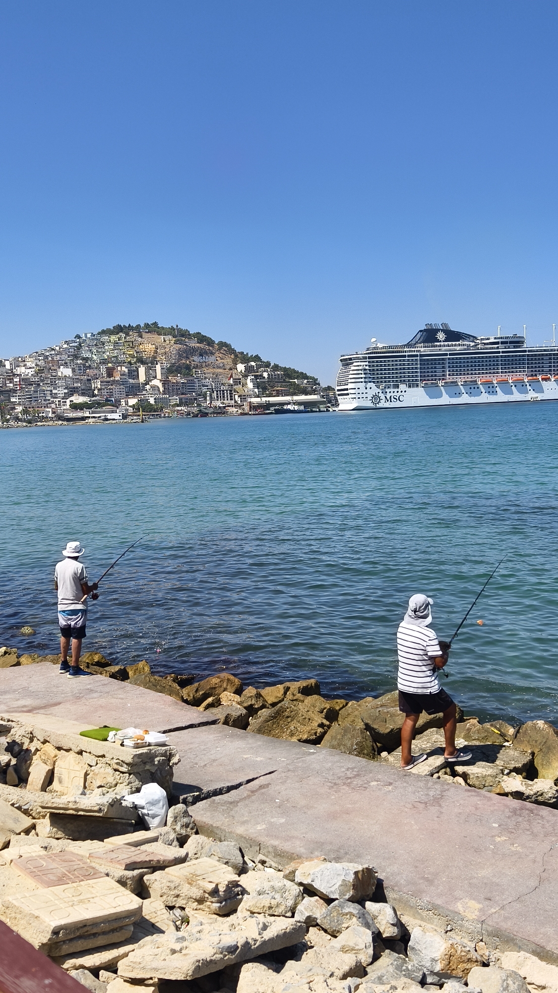 This is a photograph of the coastline in Kuşadası, Turkey, featuring two individuals fishing near the water's edge. The sky is a clear, bright blue. In the background, a large white cruise ship with the "MSC" logo is docked, and behind it, the city of Kuşadası sprawls across a hilly landscape. The foreground shows a concrete walkway and rocky shoreline.
The first fisherman, on the left, is wearing a white hat, a light-colored t-shirt, and patterned shorts. He is standing on the concrete walkway and casting a fishing line into the water. The second fisherman, on the right, is wearing a hooded cap, a striped t-shirt, and dark shorts. He is also on the walkway, facing the water and holding a fishing rod. A small orange object, possibly bait, is visible on his fishing line.
On the concrete walkway near the first fisherman, there is a green mat with what appears to be food items, including bread and other packaged goods. Scattered rocks and debris are present along the shore. The water is calm and turquoise.