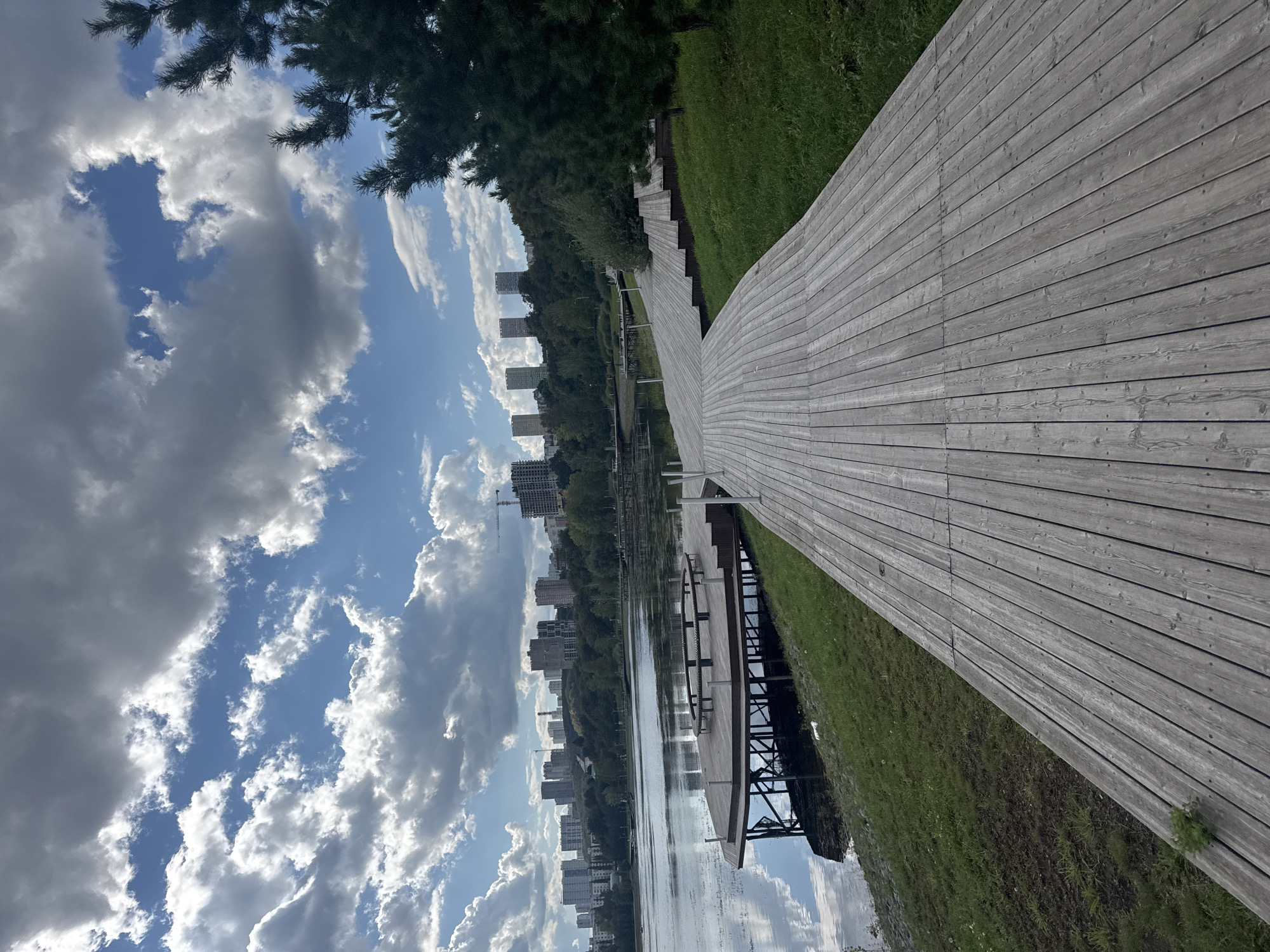 The image depicts a panoramic view of the "Arboria" park in Ufa, Russia, on a cloudy day. The foreground is dominated by a light-colored wooden promenade that curves gently to the right, with its planks running parallel to the camera's perspective. To the left of the promenade, a body of water, likely a lake, reflects the sky and the surrounding landscape. On the water, a circular wooden pier with benches is visible. The middle ground shows a grassy embankment leading up to a line of trees, beyond which a cityscape with numerous high-rise buildings and construction cranes can be seen. The sky is filled with dramatic cumulus clouds, interspersed with patches of blue. A large pine tree is partially visible on the far right of the frame.