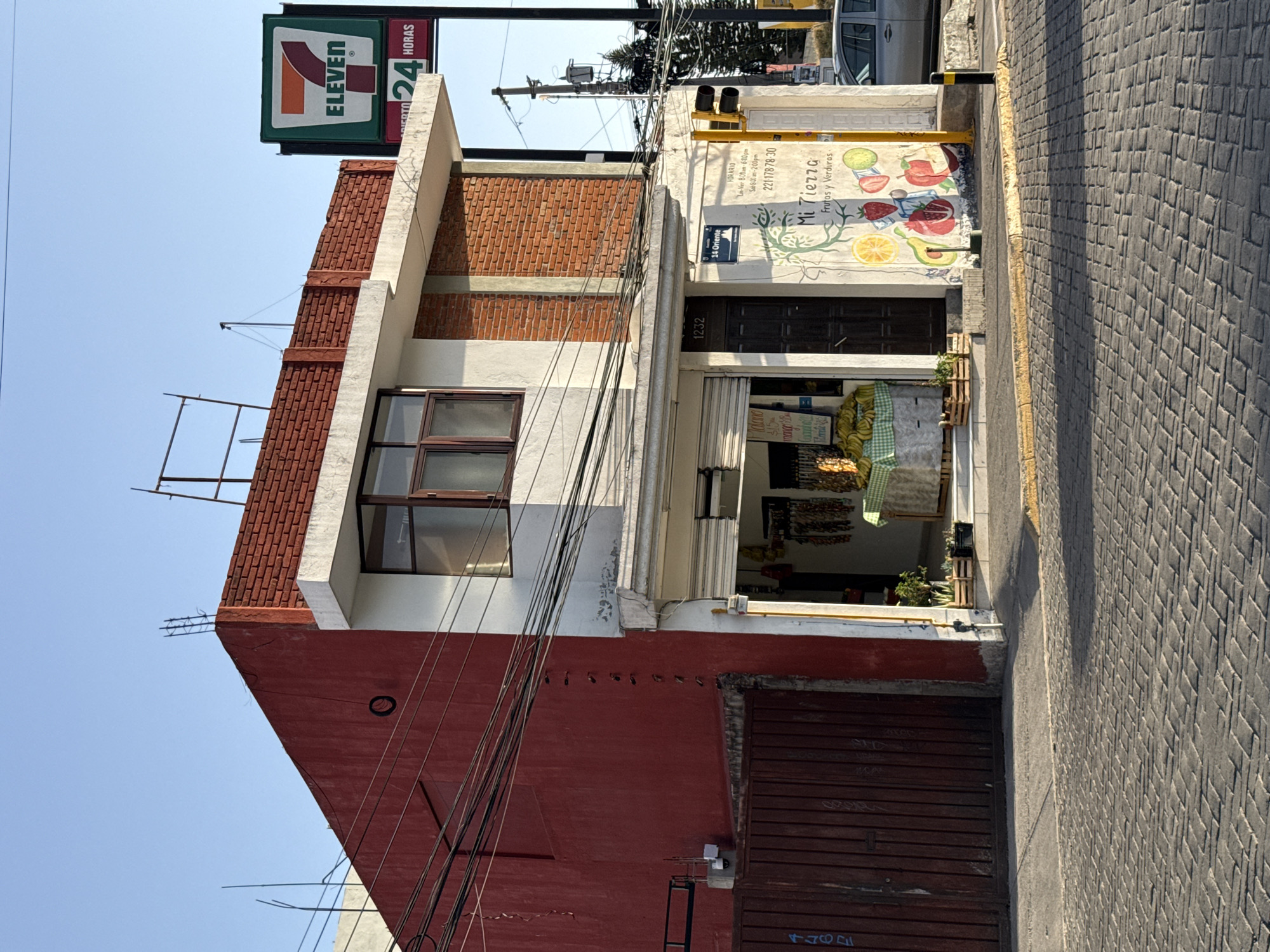 The image shows a street view in San Andrés Cholula, Mexico. The building in focus has a red and white facade, with a small store on the ground floor selling fruits and vegetables. A sign on the store wall reads "Mi Tierra Frutas y Verduras," indicating it's a local fruit and vegetable shop. The address number "1232" is visible above the store entrance, and a street sign reads "14 Oriente". The street surface is paved with textured, interlocking blocks. A 7-Eleven sign is visible above, indicating the presence of a convenience store nearby. Power lines are strung across the front of the building, creating visual clutter. A partially closed red garage door is below the fruit and vegetable store, it appears to have graffiti on it. The sky is clear blue, suggesting a sunny day.