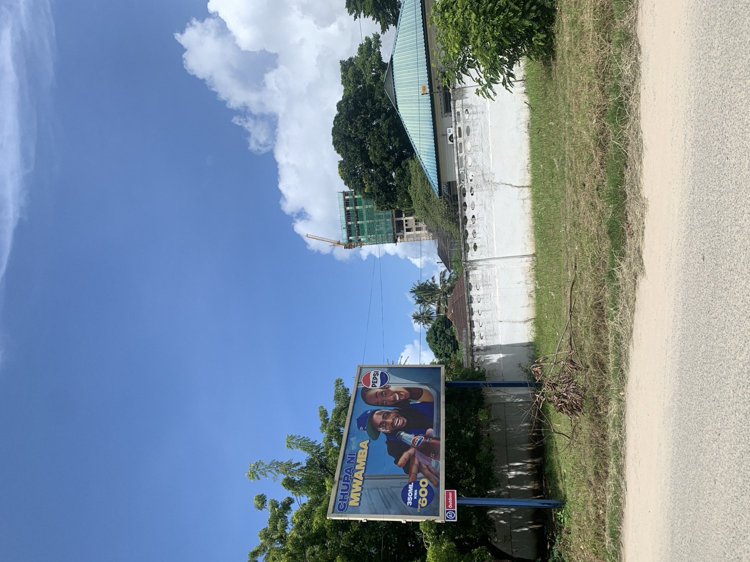 This image captures a bright, sunny outdoor scene, likely a roadside in Dar es-Salaam, Tanzania. In the foreground, a large billboard features two smiling individuals, a man and a woman, happily holding Pepsi bottles. The billboard prominently displays the "PEPSI" logo, the Swahili phrase "CHUPANI MWAMBA," and pricing information "350ML PEKEE 600," alongside an "Outdoor" advertisement logo.
The billboard is flanked by green trees and bushes. To its right, a strip of dry grass and dirt separates it from a paved road or path. Behind this vegetation lies a low, white-painted concrete wall, showing some cracks and decorated with a series of round holes.
In the background, under a clear blue sky with scattered white clouds, several structures are visible. A building with a green roof is partially obscured by trees, and further back, a multi-story building under construction, characterized by its exposed framework and scaffolding, stands next to a tall yellow construction crane. Distant palm trees also dot the horizon. The bright light and clear sky indicate it is daytime. The overall setting combines elements of urban development with natural vegetation.