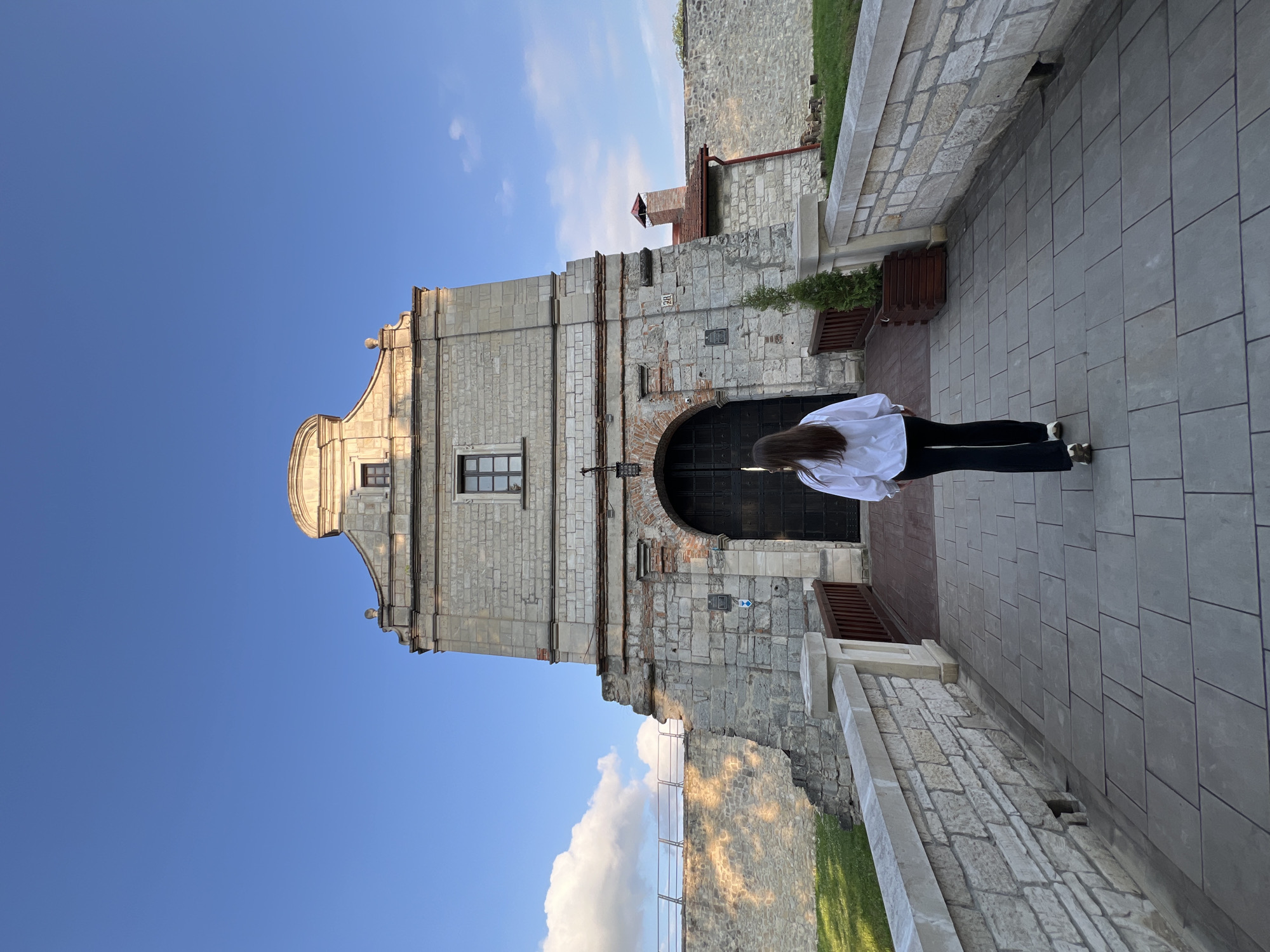 A person stands with their back to the camera, facing a large, historic stone building under a clear blue sky in what appears to be Zbarazh, Ukraine. The building, likely a castle gatehouse, is constructed from light-colored stone blocks with some sections of rougher, darker stone. It features an ornate gable at the top with a rounded pediment, two small windows, and a larger rectangular window below. A prominent, dark arched doorway serves as the main entrance, flanked by a small blue sign with a white arrow pointing right. To the right of the entrance, a wooden bench or planter is visible.
The ground where the person stands is paved with light grey rectangular tiles. To the left, a rough stone wall with grass atop it runs alongside the path. The person has long dark hair and is wearing a white long-sleeved shirt, black trousers, and dark shoes with white details. The bright, low-angle sunlight suggests either early morning or late afternoon, creating clear shadows and highlighting architectural details with a warm glow. The sky is bright blue with a few scattered white clouds. No other discernible text is visible apart from the directional arrow.