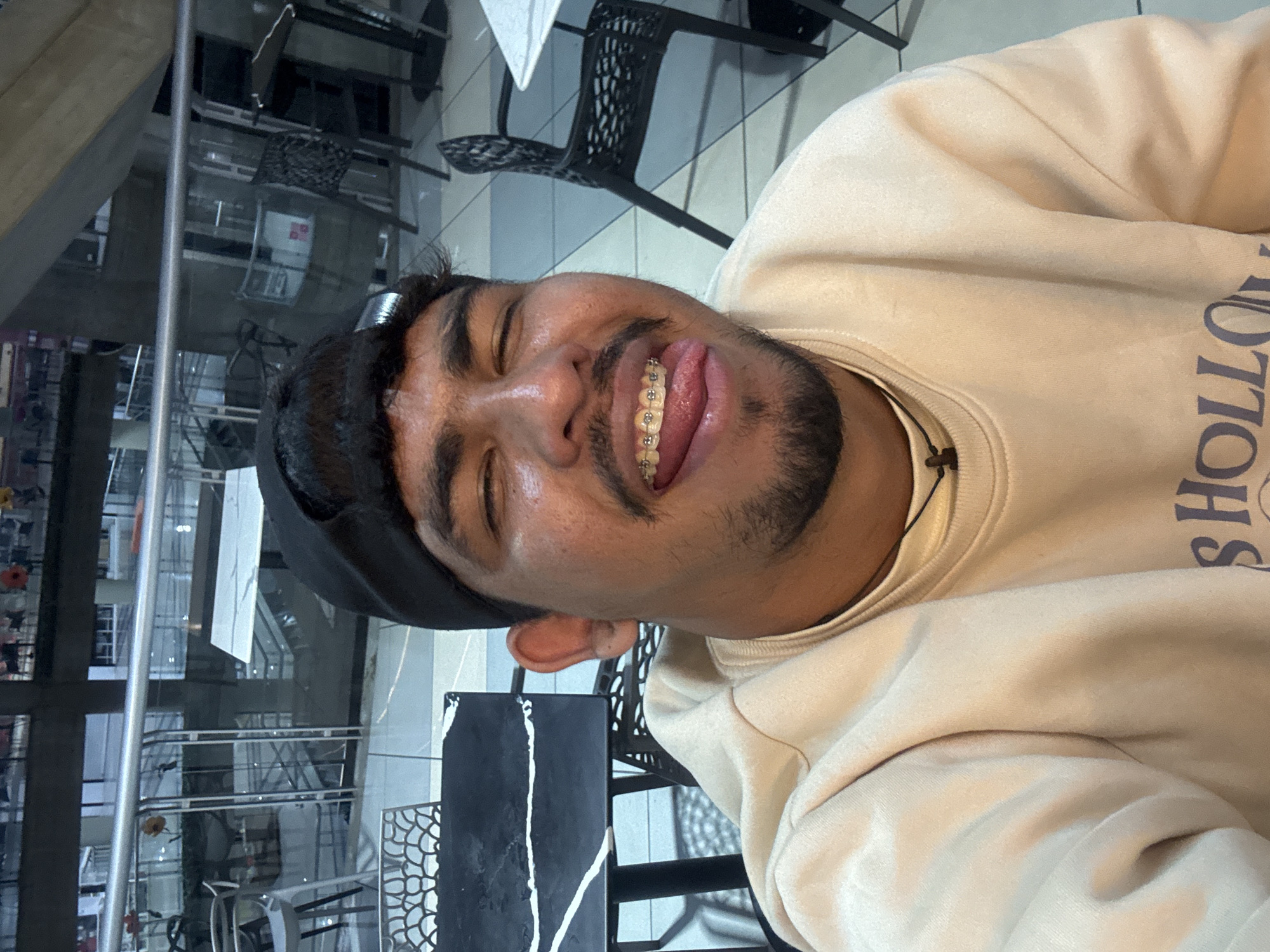 A person is posing for a selfie indoors. The person is wearing a beige sweater, a black cap, and has braces. The background shows a multi-story interior with metal railings and tables with chairs, suggesting a restaurant or similar venue. The location is plausibly Caracas, Venezuela, given the environment depicted.