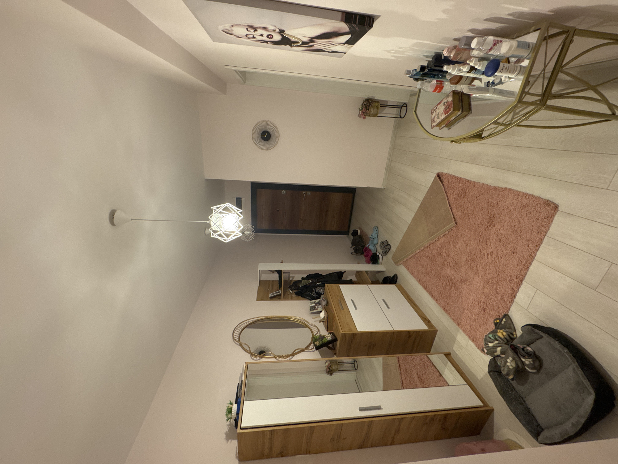 This image depicts an interior space, possibly a hallway or entryway, featuring a combination of furniture and decorative elements. The color palette leans towards light tones, with pink-beige walls. Furniture includes a wooden cabinet with white drawers, a mirrored dresser, and a decorative gold shelf unit holding toiletries. A light fixture hangs from the ceiling, and a round wall clock is visible. A pink rug and a pet bed occupy the floor space. A framed image of Marilyn Monroe is affixed to the ceiling. It's not possible to determine the specific location in Burgas, Bulgaria, from this image alone.