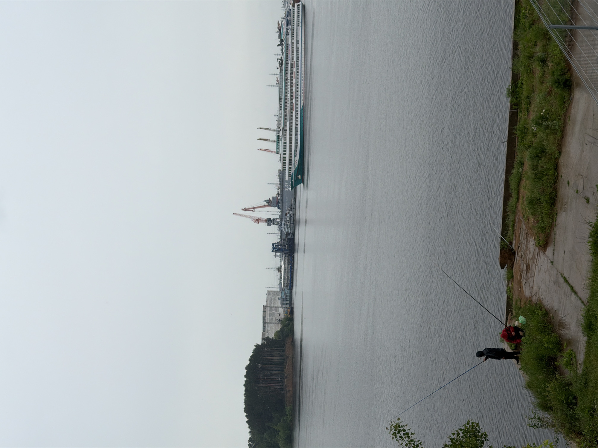 This image, taken in Chaikovsky, Russia, depicts a wide river scene under an overcast sky, creating a muted, grey palette across the water.
On the left bank, a long, multi-deck white cruise ship, featuring teal accents on its hull, is prominently docked. Adjacent to the ship is an industrial port characterized by several large cranes, some red and some grey, and various buildings. Further along this bank, a dense forest of evergreen trees lines the water's edge.
The right bank presents a contrasting view with a concrete path running alongside the river, bordered by green grass and bushes. A metal fence is visible towards the top of this bank. Two individuals are engaged in fishing from this side. One person, dressed in dark attire, stands closer to the viewer with a fishing rod cast into the water. Further back and partially obscured by foliage, another person in a red jacket is seen, likely also fishing.
The time of day appears to be daytime, though the overcast conditions provide soft, diffused lighting. The scene combines natural elements like the river, forest, and grass with industrial infrastructure and recreational activity. Faint text reading "SLOW" is visible on the hull of the cruise ship, followed by another less discernible word.