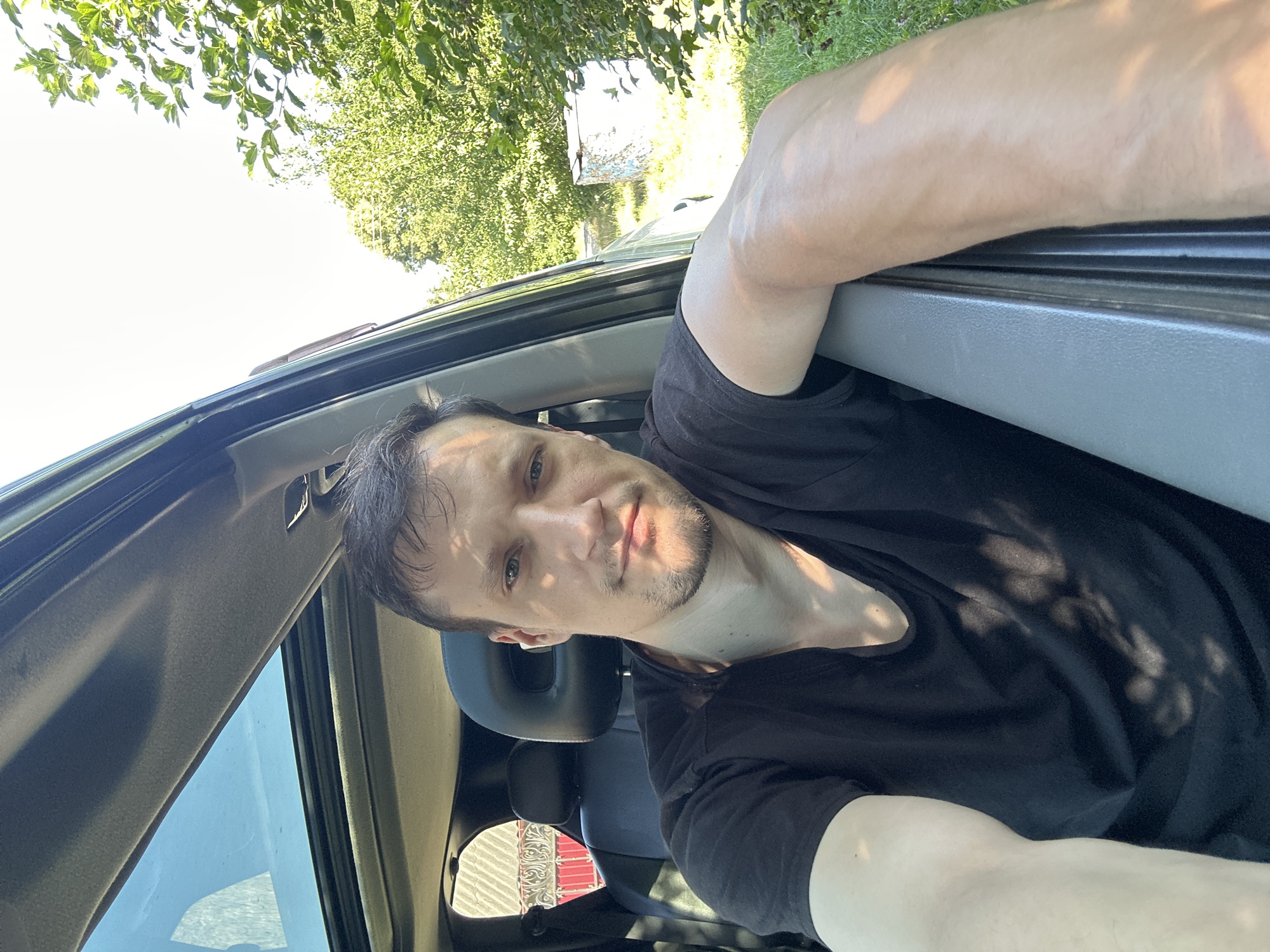 In Shatava, Ukraine, a man is pictured inside a vehicle, resting his arm on the open door frame. He is wearing a black t-shirt, and the vehicle's sunroof is open, revealing a clear sky. The background shows greenery, indicating an outdoor setting. The image is taken during daylight hours, with sunlight casting shadows on the man and the car's interior.