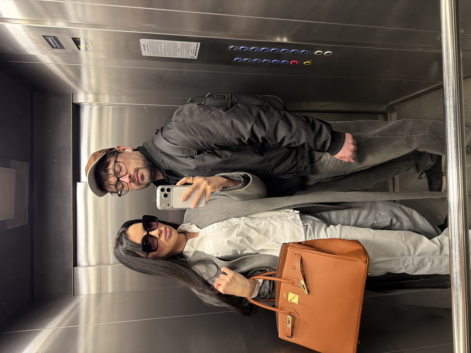 Inside a brushed stainless steel elevator in Moscow, Russia, a female and a male stand, captured in a selfie reflection. The female, positioned on the left, has long dark hair, wears large dark square-framed sunglasses, a white collared shirt, light grey trousers, and a long grey coat. She holds a silver smartphone with three camera lenses in her right hand to take the photograph and carries a large, structured light brown leather handbag with gold-tone hardware. Her left hand is near the bag's strap. She wears a black choker necklace and multiple rings. The male, standing slightly behind and to her right, wears a brown baseball cap, clear-framed glasses, and has white wireless earbuds in both ears. He is dressed in a black bomber jacket over a dark shirt, dark grey jeans, and black sneakers. A black backpack is visible on his back. Both individuals are looking directly towards the camera. The elevator's right wall features a control panel with illuminated circular buttons, including a red "0", a yellow button, and blue buttons for floors "1" through "4". Above the buttons, a digital display shows "3". A reflective label on the right wall displays Cyrillic text, including "ООО «ТЕНКОМ»" (TENCOM LLC) and a telephone number with a "8 (495)" area code, confirming the location in Moscow.