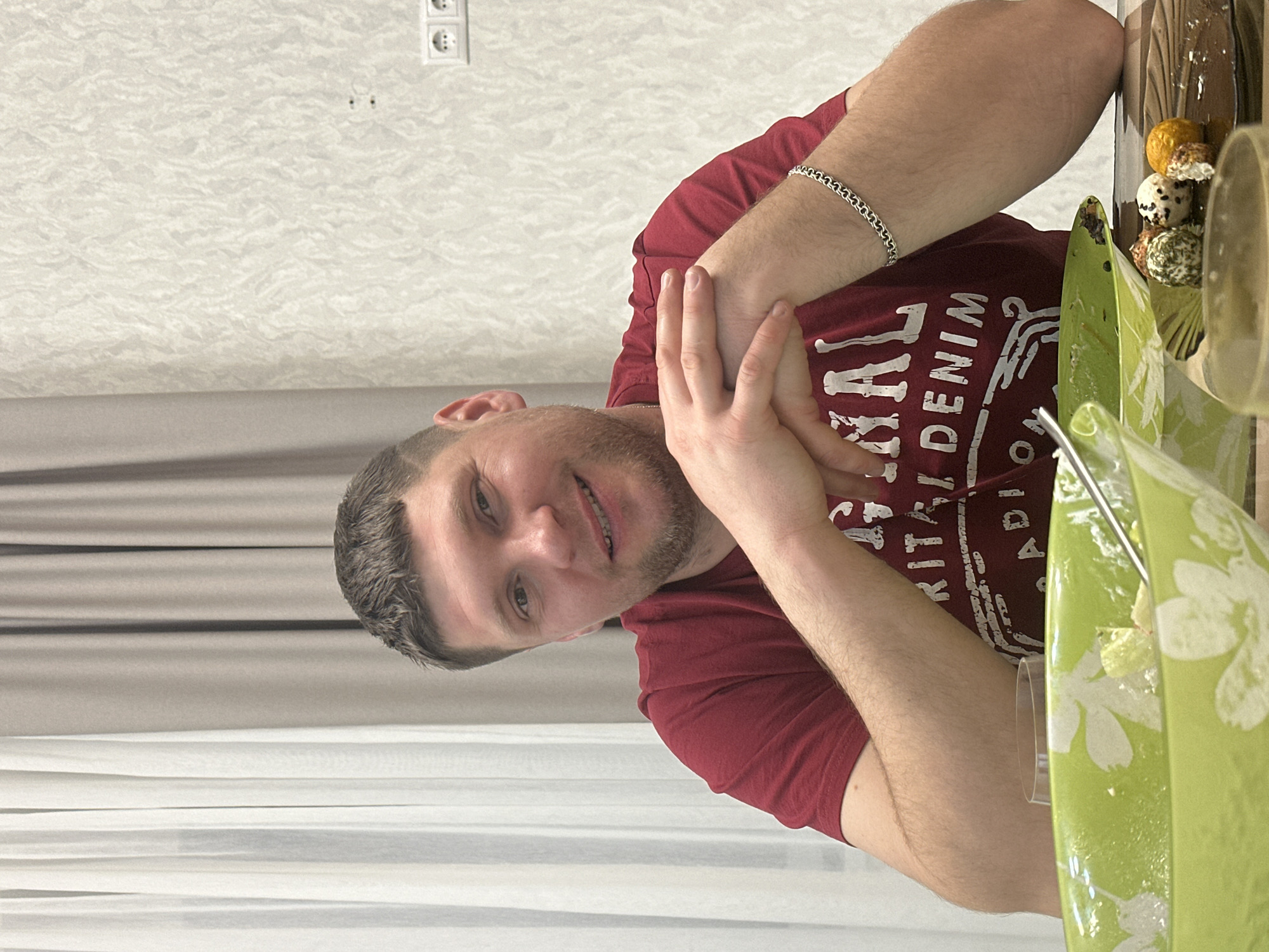A man wearing a maroon t-shirt and a silver bracelet is seated at a table with food. The food includes decorated balls of cheese. A green serving dish with white flowers is also on the table. Behind him are light-colored curtains, and textured wallpaper is visible above the curtains. Electrical outlets are mounted on the wall. The location is presumed to be in the city of Sloboda Staraya, Russia.