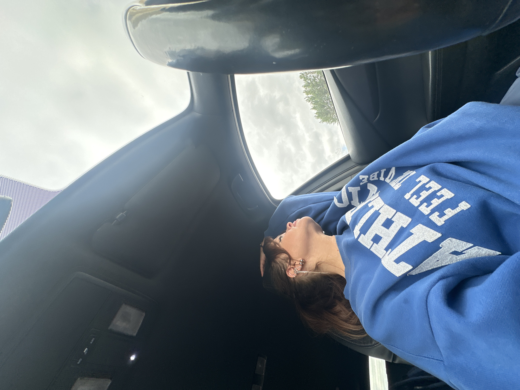 A woman with brown hair in a ponytail, wearing a blue sweatshirt with white lettering, is sitting in the passenger seat of a car. She is looking out the window and has her hand on her head. The car has a sunroof, which is open. The sky is cloudy and there are trees visible outside the car. There are no specific indicators in the image that place the location in Chaikovsky, Russia.