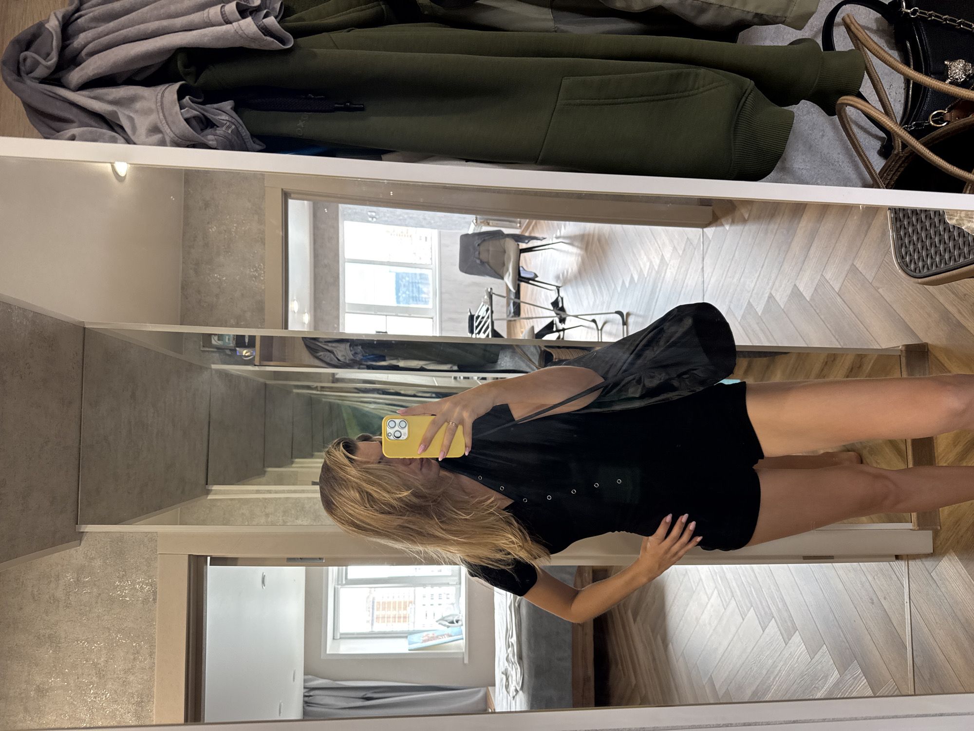 A woman is standing in a room, reflected in a large mirror. She is wearing a black romper and holding a yellow phone. The room appears to be a dressing area or hallway, with a bed visible in the background through a doorway on the left, and coats hanging on the right side of the mirror. The flooring is a herringbone pattern of light wood. The overall lighting is bright and natural, suggesting daytime.